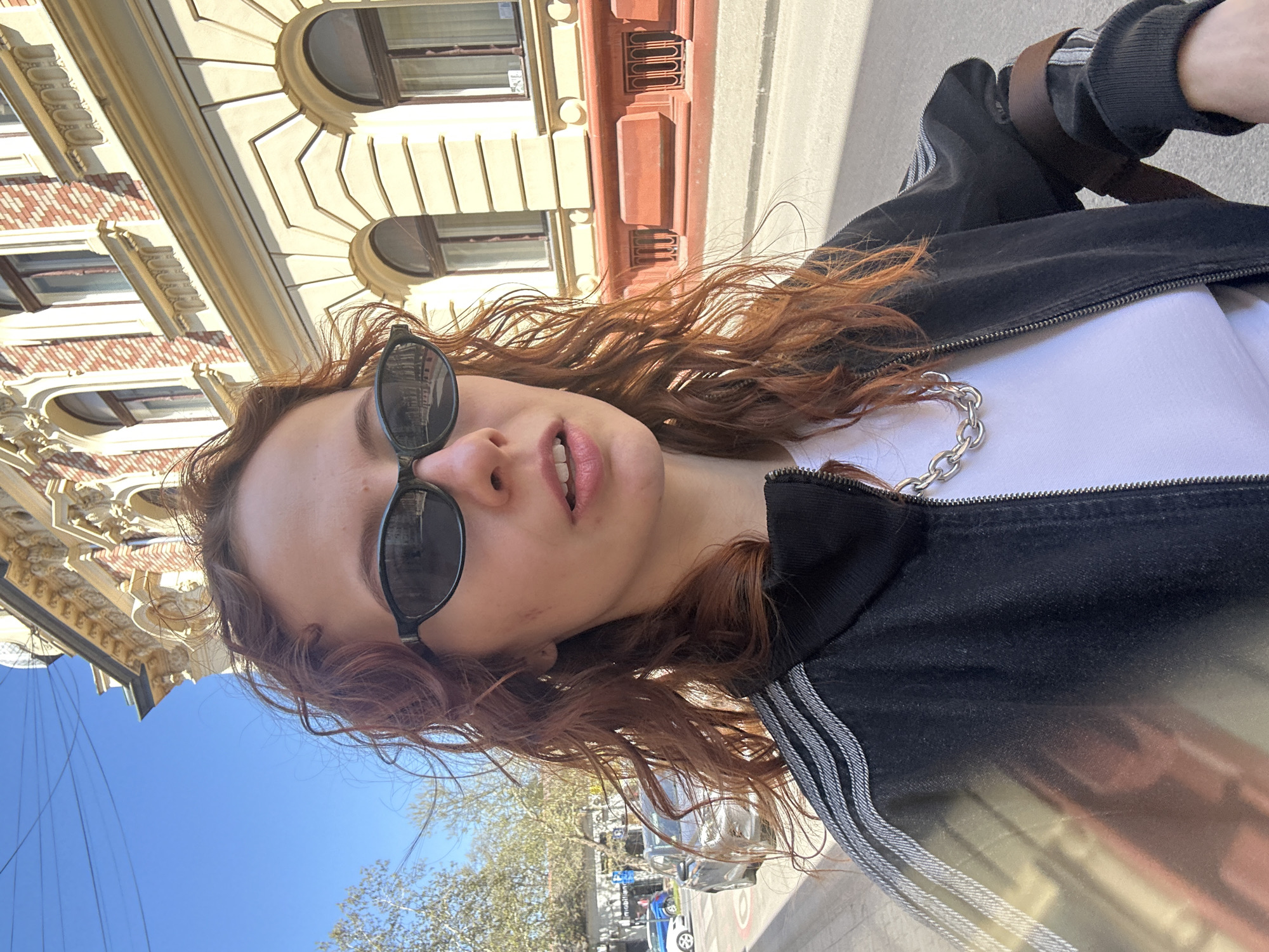 A young woman with curly, reddish-brown hair is wearing sunglasses, a white shirt, and a black jacket with three white stripes on the shoulder. A silver chain is visible around her neck. She is standing on a street in Novi Sad, Serbia, as suggested by the architecture in the background. The architecture includes decorative, multi-story buildings with windows and ornate facades. The weather appears sunny. Cars and trees are visible in the background street scene.