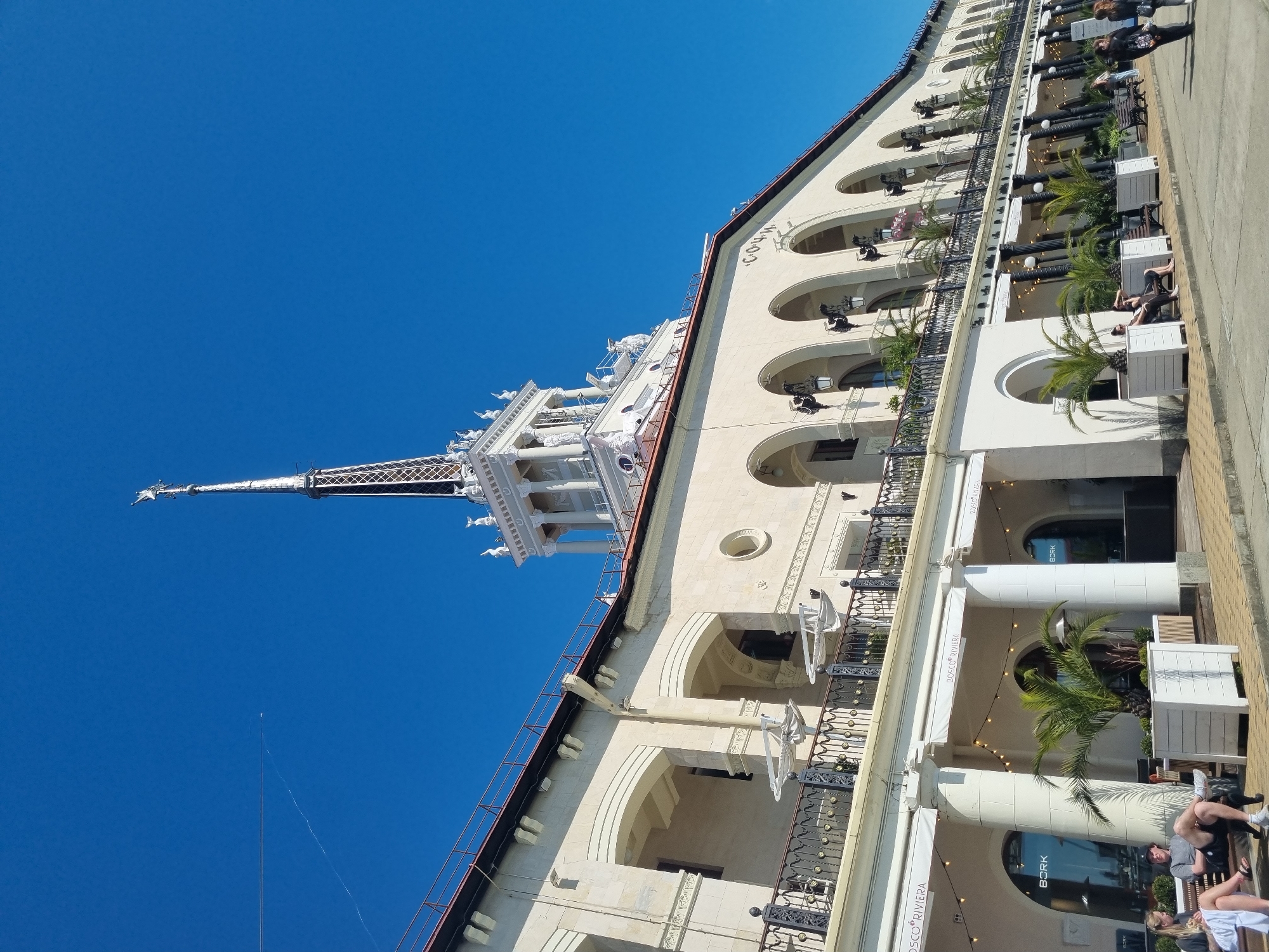 The image presents a low-angle view of a building with white architecture against a clear blue sky. The building features a tall spire, several arched windows, and decorative architectural elements. A section of the building's facade is visible on the right, showing an area with columns, arched entrances, and what appears to be outdoor seating with potted palm trees. People are seated at some of the tables. The image has been rotated, giving the building an angled appearance. The location appears to be in Sochi, Russia.