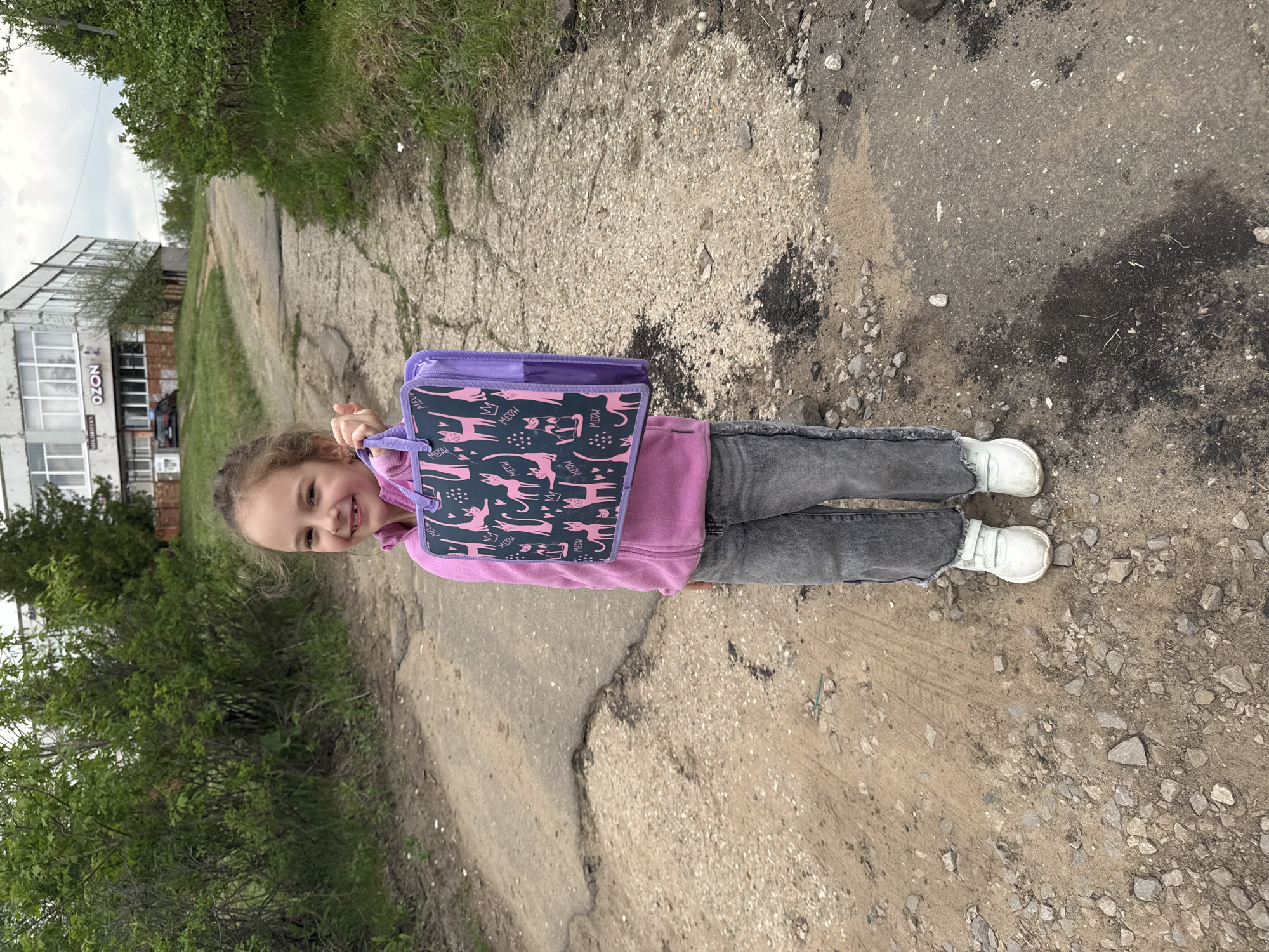 A young girl stands on a cracked asphalt road with green foliage on either side. She is wearing a pink hooded sweatshirt, gray jeans, and white shoes. She is carrying a purple backpack with pink cat designs. A building with a sign that says "OZON" is visible in the background. The location is Shevlyakovo, Russia.