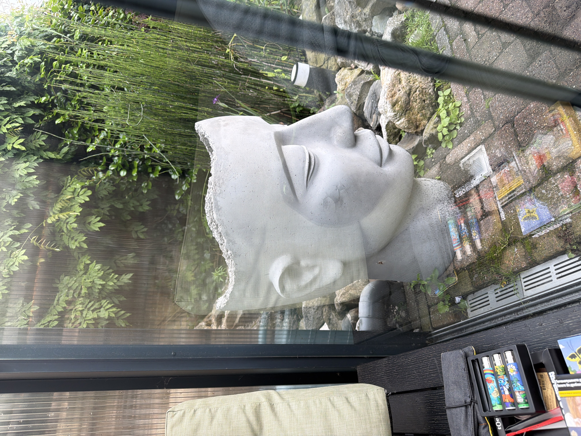 The image shows a statue of a head, likely made of concrete, with a partially broken or open top. The statue is positioned in a garden area visible through a glass panel, possibly a window or greenhouse. Behind the statue, there are tall, thin green stalks of plants, resembling horsetail or bamboo, and rocks of various sizes. Further in the background, there is dense green foliage. In the foreground, on a dark wooden surface, a black case contains several colorful cylindrical objects, possibly vape pens or similar items, arranged in a tray. The ground appears to be paved with brick-like stones. The overall scene suggests an outdoor setting, potentially a backyard or garden space, with elements of decorative sculpture and natural vegetation. There is no indication of the specific location in Tilburg, Netherlands.
