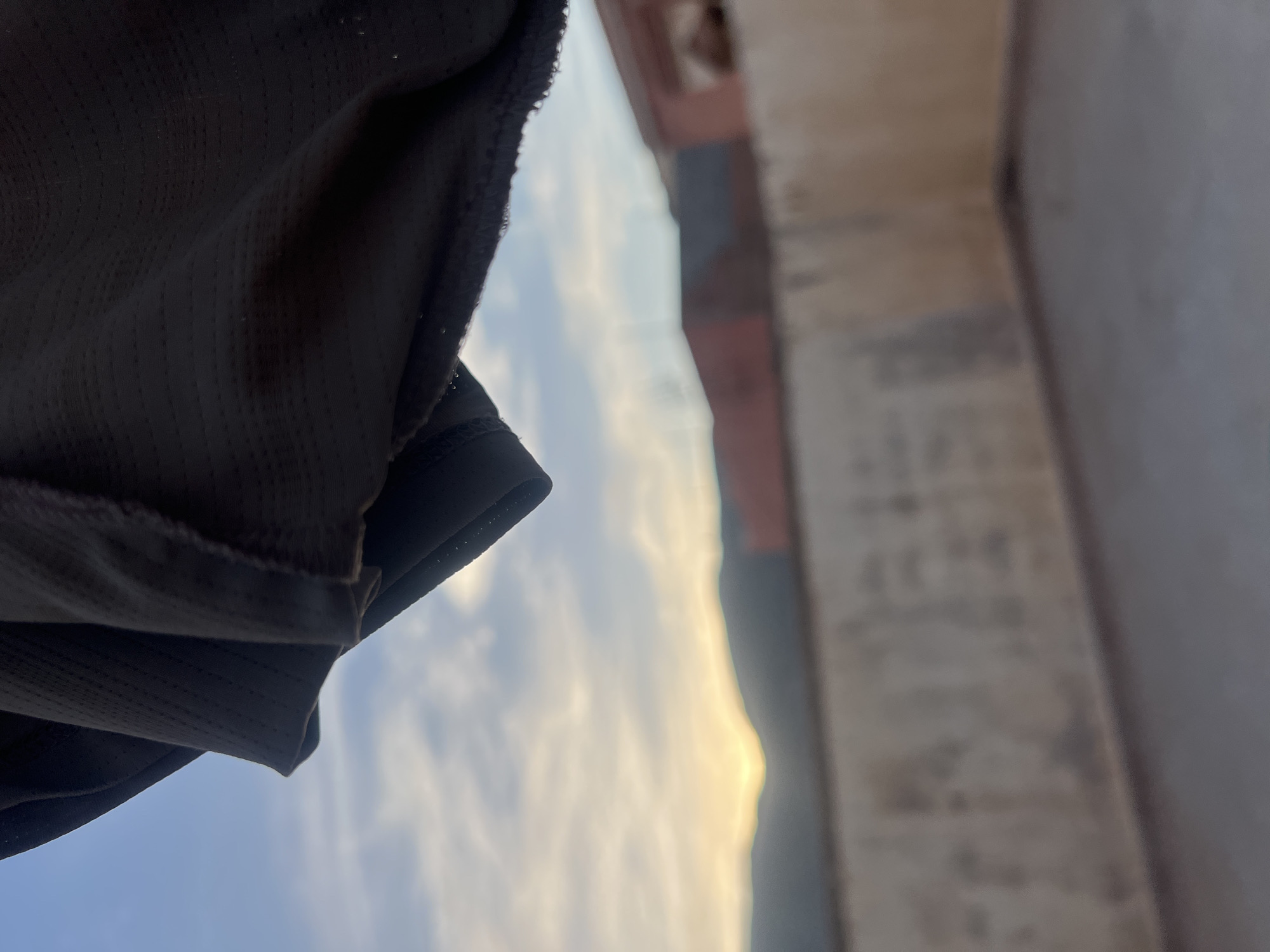 The image shows a view from what appears to be a building in Ouaouizert, Morocco. A portion of dark fabric is visible in the foreground on the left. A blurred building or wall is visible on the right. In the background, the sky is visible with some clouds. There are also what appear to be distant hills or mountains, with a bright glow suggesting the sun near the horizon. The image is shot from a low angle, looking upward.