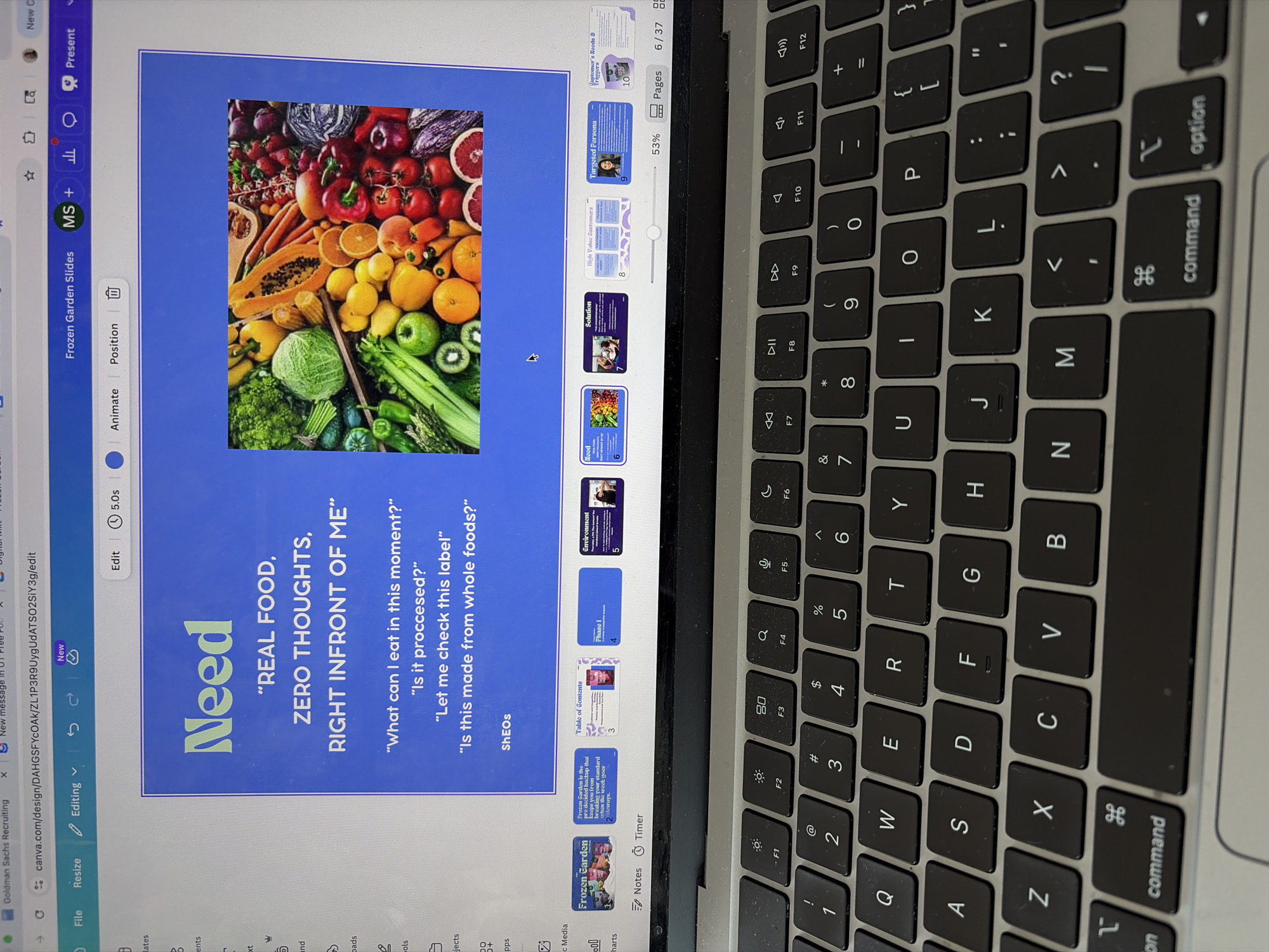 The image captures a close-up, angled view of a person operating a MacBook laptop, displaying a Canva presentation. The scene is an indoor setting, likely a workspace, with the keyboard in the foreground.
The primary subject is a digital presentation open on the laptop screen. The current slide, numbered 6 out of 37, is titled "Need" and features a large, colorful image of various fresh fruits and vegetables, including papaya, oranges, kiwis, cabbage, bell peppers, tomatoes, and leafy greens. Overlaying this image and on the blue background to the right, prominent text reads: "REAL FOOD, ZERO THOUGHTS, RIGHT INFRONT OF ME". Below this, several questions are listed: "What can I eat in this moment?", "Is it proccesed?", "Let me check this label", and "Is this made from whole foods?". A small text "ShEOs" is at the bottom.
On the right side of the screen, a sidebar displays thumbnails of other slides in the presentation. Visible titles include "Frozen Garden", "Frozen Garden is the pre-decided backup...", "Table of Contents", "Phase I", "Environment", "Solution", "High Value Customers", "Targeted Persona", and "Customer's Needs & Triggers". The Canva interface shows "Frozen Garden Slides" as the presentation title, with editing options like "Edit", "Animate", "Position", and "Present". The browser URL indicates "canva.com/design...", confirming the platform. Other visible text from the browser tabs includes "Goldman Sachs Recruiting" and "New message in UI Free Pub".
The laptop's keyboard, primarily the right half, is visible in the foreground, showing standard keys such as letters (QWERTY, ASDFGHJ, ZXCVBNM), numbers (1-0), and function keys (F1-F12), along with "command" and "option" keys. The image provides no specific cues for the time of day or emotions, but the context provided is Austin, United States of America.