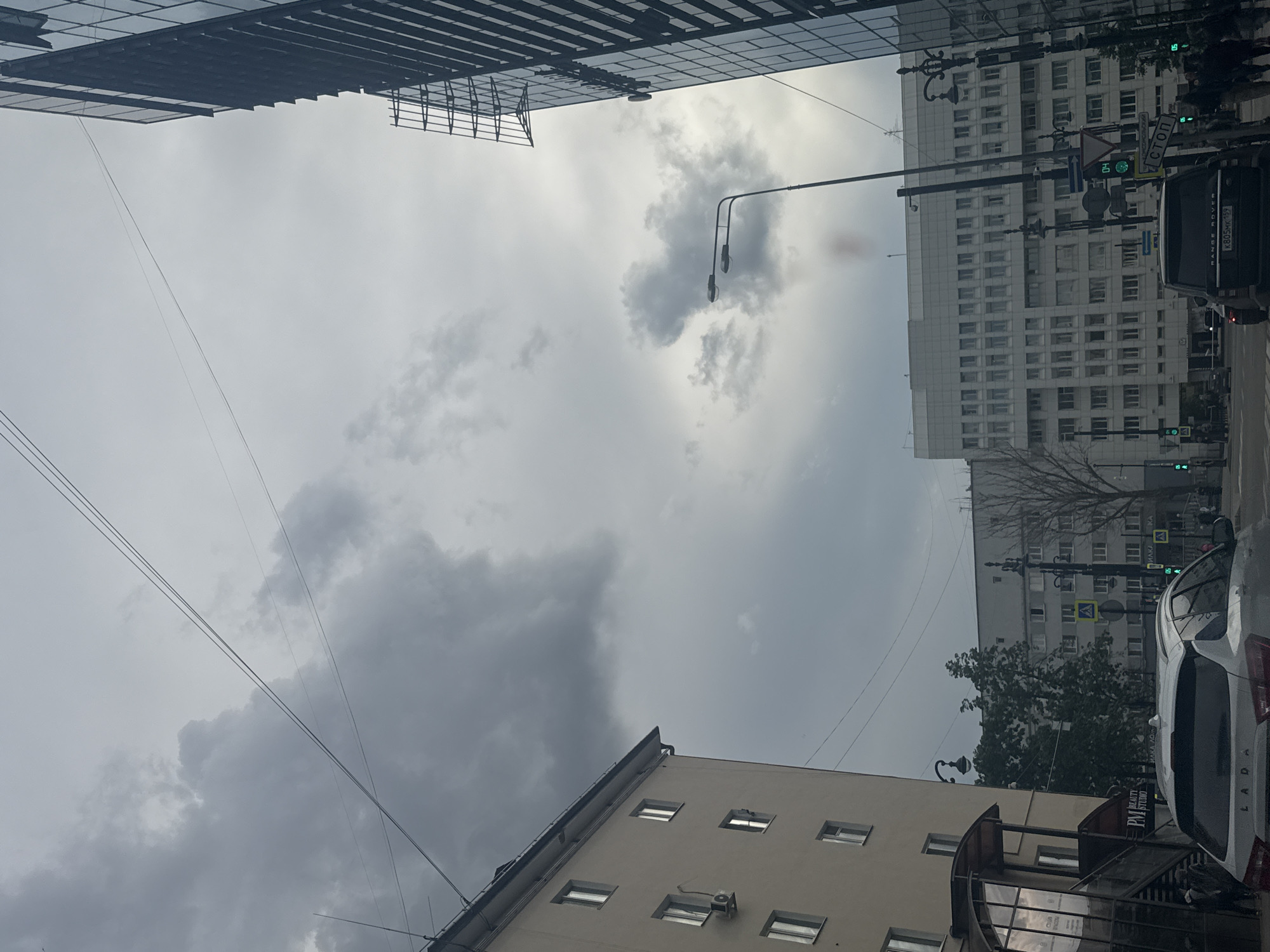 This media file depicts an urban street scene in Perm, Russia, under a cloudy sky. The perspective is from inside a vehicle, looking upwards and outwards. The scene is characterized by overcast weather, with gray clouds dominating the sky.
Prominently featured are several multi-story buildings. One large, modern building with a glass facade and dark horizontal elements is visible at the top. To its right, a white, blocky building with many windows occupies a significant portion of the frame. Further down the street, another building with a lighter facade and multiple windows is visible. A smaller building with a beige exterior and a dark, arched entrance is in the foreground on the right.
Street elements include traffic lights showing green signals, street lamps, and traffic signs, including one that reads "СТОП" (STOP). A dark-colored Range Rover is parked or moving in the street, and a white Lada is partially visible in the foreground on the right. Power lines crisscross the sky, adding to the urban feel. The overall impression is that of a typical day in a Russian city, with the weather contributing to a somewhat subdued or muted atmosphere. The time of day appears to be daytime, given the ambient light, but the overcast conditions obscure any distinct shadows that might indicate a precise hour. There are no people clearly visible in the frame.