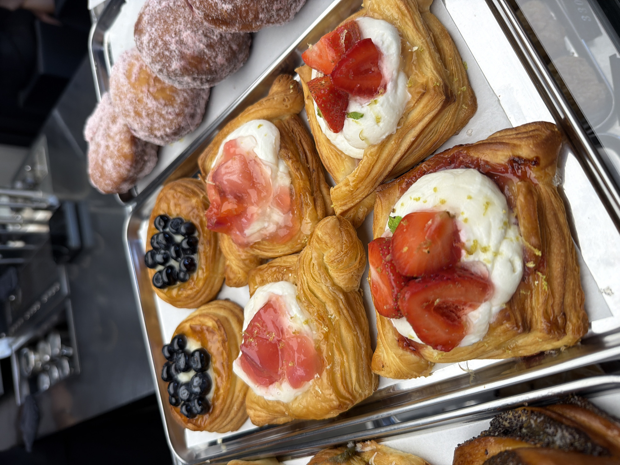A close-up, eye-level shot shows a tray filled with assorted pastries, likely in a bakery or cafe setting. The pastries are arranged on a metal tray lined with white paper. In the foreground, three square danishes with flaky, golden-brown pastry are visible. The leftmost danish is topped with a dollop of white cream, glistening strawberry jam, and a sprinkle of finely grated zest. To its right, another danish features a similar cream and jam topping, with a few fresh strawberry slices and a mint leaf. A third danish, partially obscured, appears to be topped with blueberries. Behind these, to the left, are two round danishes, each topped with a generous amount of blueberries. Further back, to the right, are three round doughnuts coated in a pinkish, powdered sugar-like substance. The background shows a blurred view of stainless steel equipment, possibly a coffee machine or counter, suggesting a commercial food preparation environment. The lighting is bright, highlighting the textures and colors of the pastries. The image does not contain any information indicating its location in Izhevsk, Russia, or any people or actions.