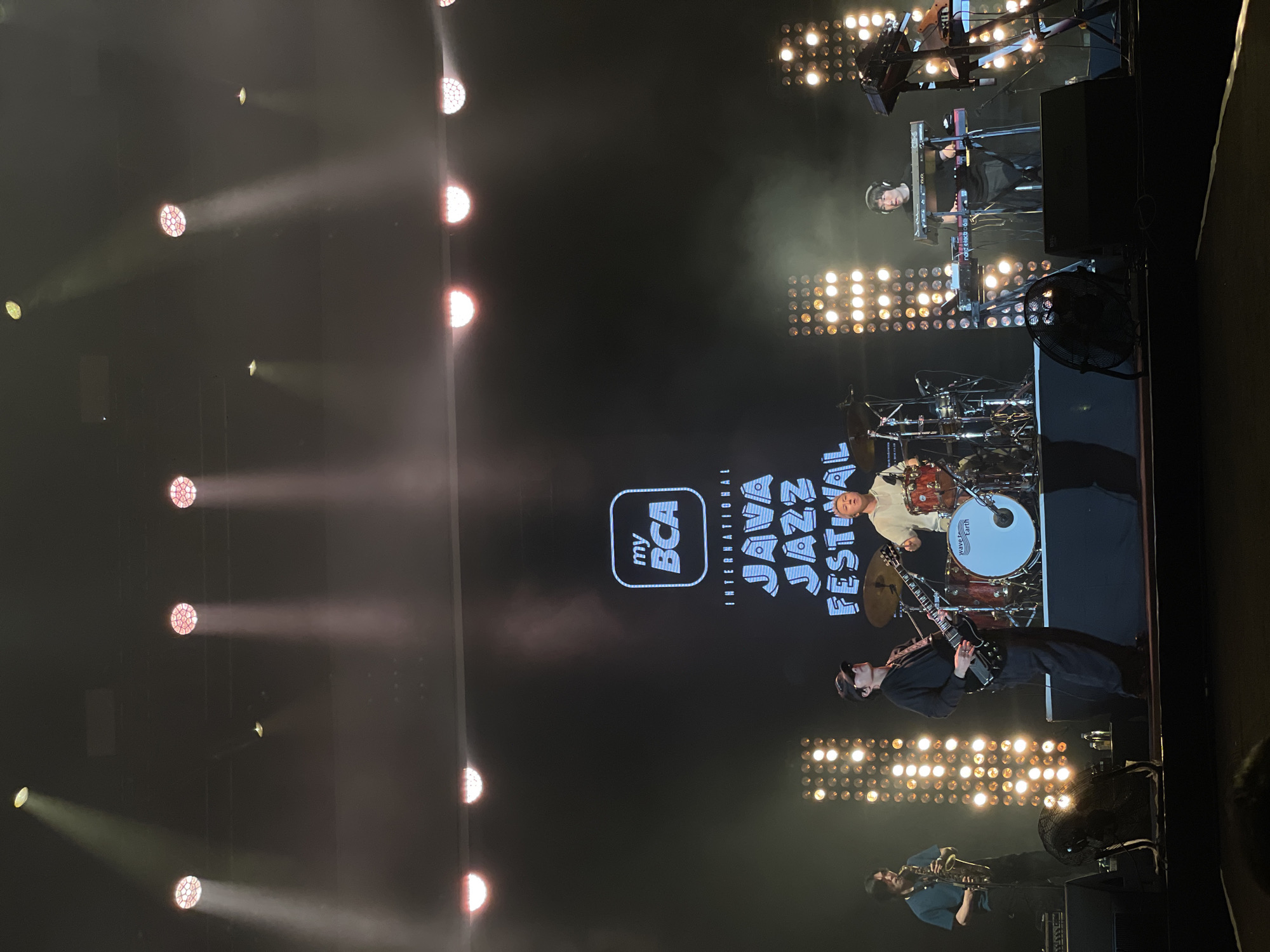 The image shows a musical performance on a stage at the "myBCA International Java Jazz Festival". The location is likely Salembaran, Indonesia, as the festival takes place there. Several musicians are visible: a drummer, a guitarist, a saxophone player, and a keyboardist. Stage lighting illuminates the performers and the festival backdrop.