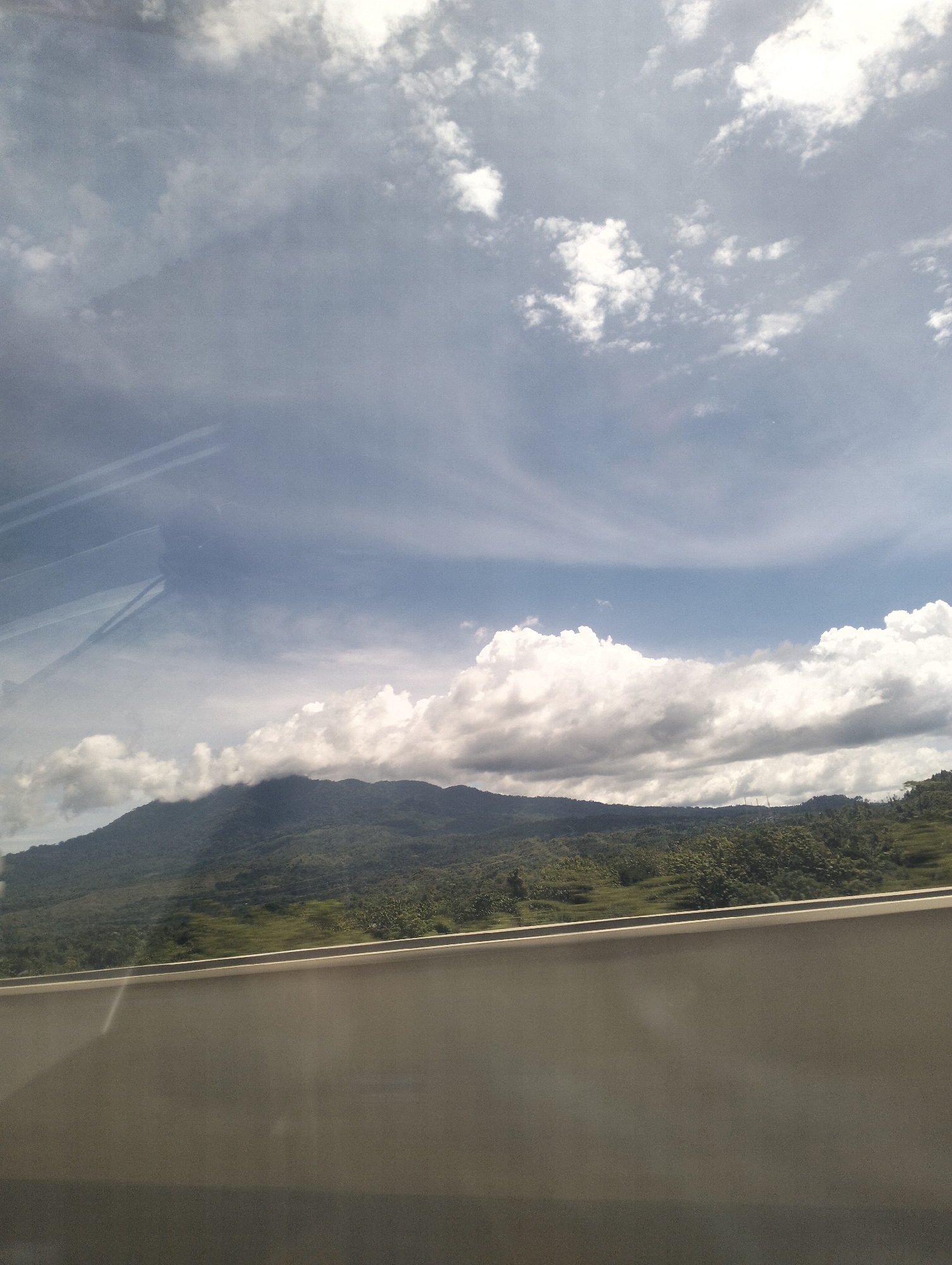 The image captures a scenic view of mountains and sky, possibly taken from a moving vehicle. The sky is filled with dramatic clouds, some of which are white and puffy, while others are darker and more diffused, hinting at potential weather changes. A range of lush green mountains dominates the middle ground, with some peaks shrouded in clouds. The foreground is occupied by a solid, light-colored structure, possibly a barrier or wall, which appears to be part of the viewpoint. The overall impression is one of natural beauty and vastness. There are no people or specific man-made structures visible that would definitively pinpoint the location to Lamtamot, Indonesia.