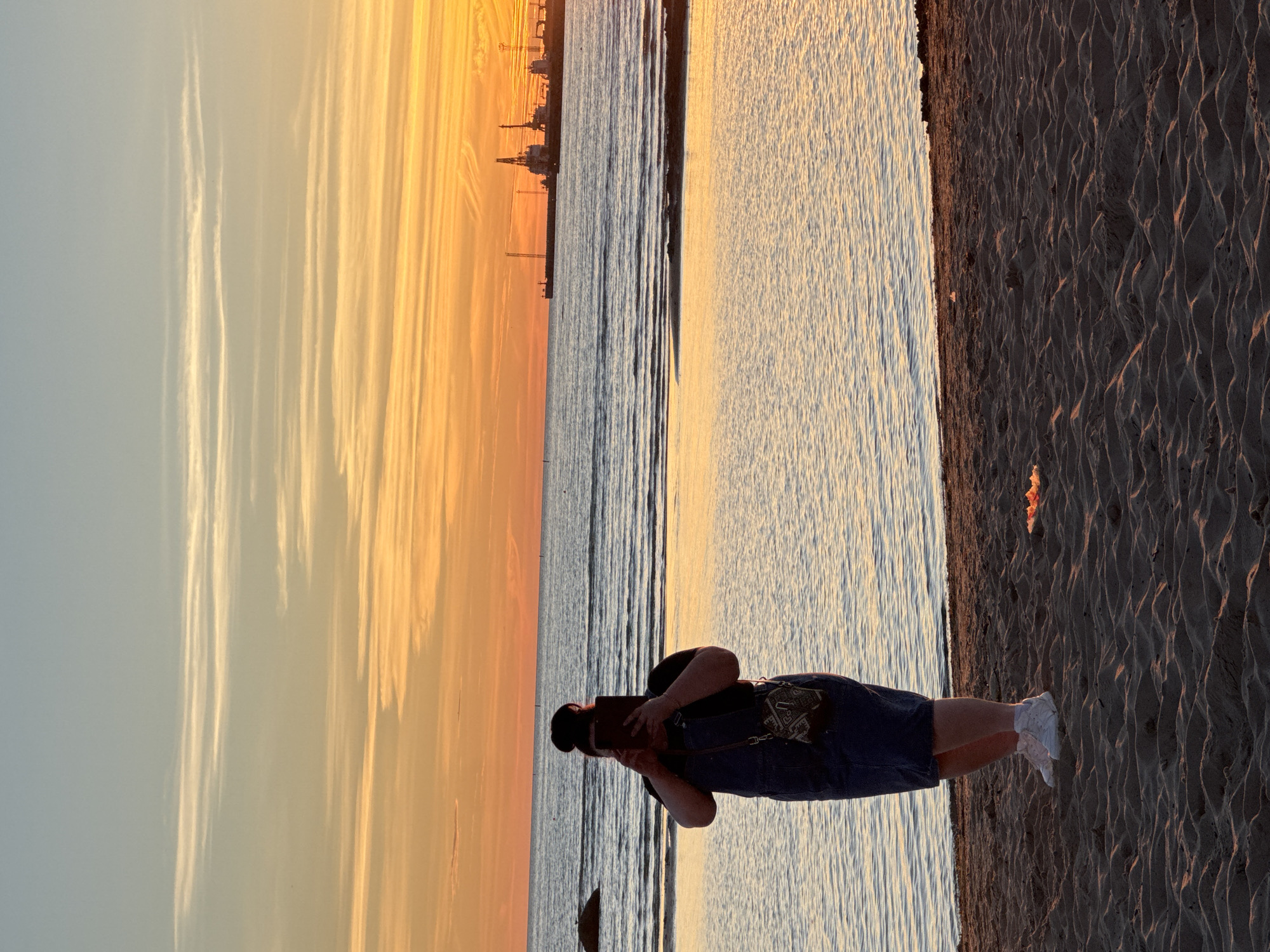 The image depicts a person standing on a sandy beach at sunset, facing the sea. The person is holding up a tablet or phone, possibly taking a photograph or video, with their face obscured by the device. They are wearing denim overalls, a black t-shirt, and white sneakers, and have their hair tied up in a bun. A patterned bag is slung across their shoulder.
The sea stretches out to the horizon, reflecting the warm hues of the sunset. In the distance, along the coastline, industrial structures including cranes and buildings are visible, suggesting an industrial port area. The sky is a vibrant blend of orange, yellow, and pale blue, with wispy clouds. The foreground is a sandy beach with ripples from the wind. This scene is consistent with the coastline near the Kalaranna area in Tallinn, Estonia, known for its beaches and proximity to the harbor.