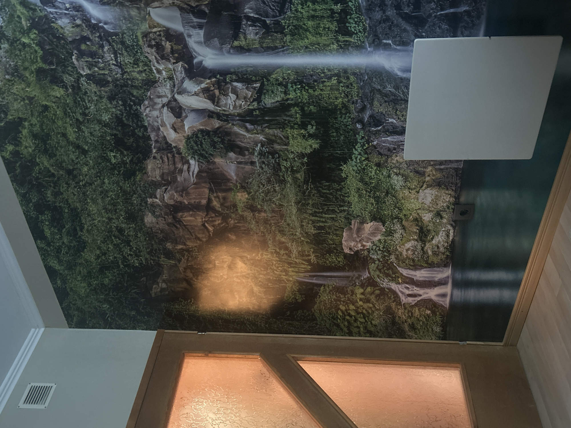 The media file captures an indoor scene dominated by a large, vivid wall mural. The mural depicts a lush, green natural landscape featuring multiple cascading waterfalls flowing over rugged, dark rocky cliffs. Abundant green foliage, including dense trees, bushes, and smaller plants, covers the rocks and surrounding areas, giving the impression of a vibrant, verdant environment.
In the upper right section of the image (as oriented), a plain, light-colored rectangular panel, possibly an acoustic ceiling tile or a large light fixture, is visible against a dark teal or green painted wall. Below this, an electrical outlet can be seen on the same wall.
The foreground is occupied by the upper part of a wooden door or a large window frame. This frame, made of light to medium brown wood, encloses textured, translucent glass panels. A prominent, bright golden-yellow light spot is cast upon the mural directly above this door/window, suggesting an interior light source. To the left of the door, a section of a light-colored wall is visible, featuring a white rectangular air vent with horizontal slats.
The image appears to be taken indoors, likely under artificial lighting. No people or visible text are present. The overall setting suggests a uniquely decorated interior space, possibly a room or hallway, with an immersive natural theme.