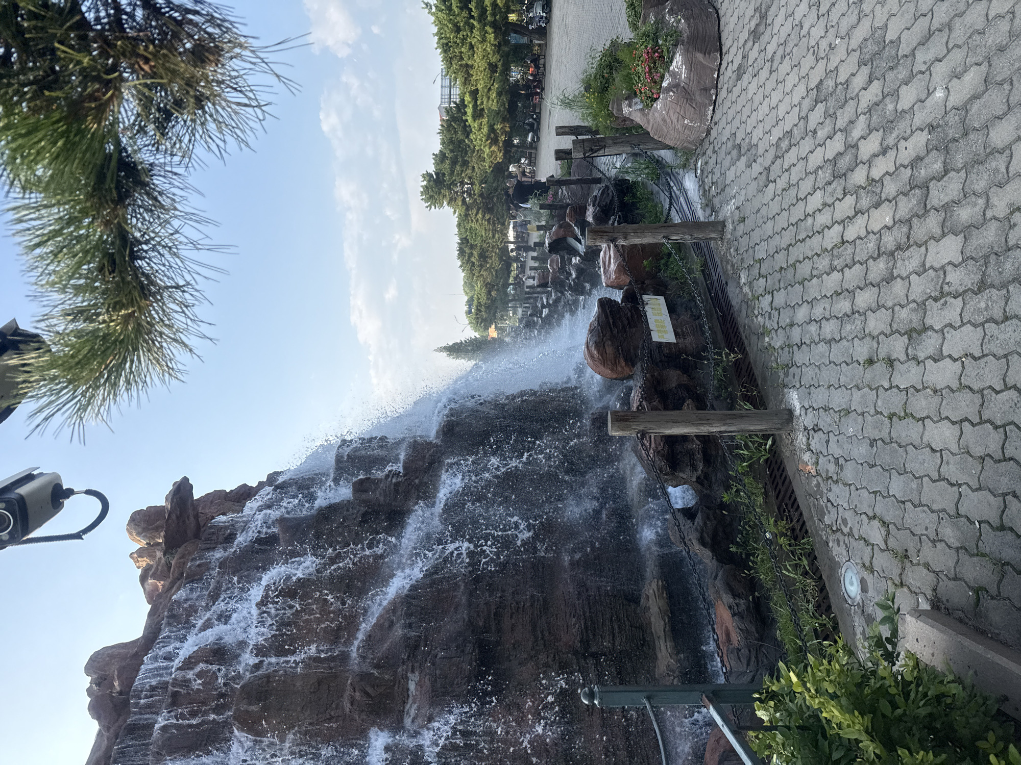The image depicts an outdoor scene featuring a man-made waterfall cascading down a rocky structure. In the foreground, a paved walkway made of interlocking gray bricks leads towards the waterfall. A green metal railing runs along the left side of the walkway, adjacent to lush green bushes. Wooden posts and chains delineate the area around the base of the waterfall, where a metal grate is visible.
Behind the waterfall, a line of trees with lush green foliage forms a backdrop, with a clear blue sky overhead. Several people are visible in the distance, walking or standing near benches, and a number of parked motorcycles and scooters are present. A surveillance camera is mounted overhead, attached by a flexible black cable. The overall environment suggests a public park or recreational area, likely in a city given the parked vehicles and distant buildings. The presence of a Chinese sign near the waterfall indicates the location is in China.
Based on visual cues within the image, such as the style of the landscaping, the type of trees, and the signage, this location can be identified as being within the **Beidaihe District of Qinhuangdao, China**. Specifically, the arrangement of the waterfall, walkway, trees, and the presence of public amenities are characteristic of areas in Beidaihe known for their scenic beauty and tourist appeal.