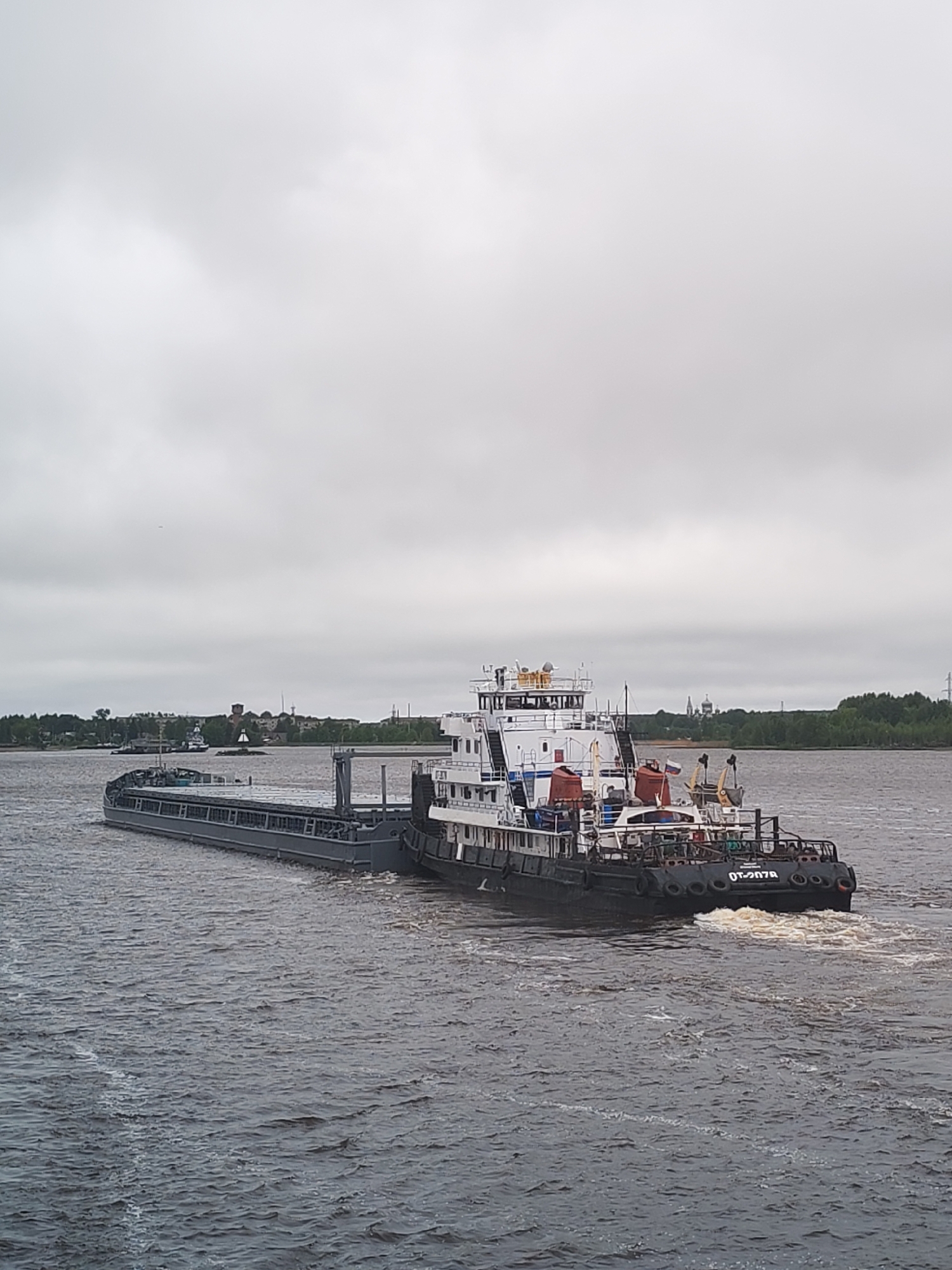A medium shot depicts a tugboat pushing a barge on a river under an overcast sky. The tugboat is black and white, with multiple decks and a visible Russian flag. The barge is long and gray. The river surface is choppy, with visible wake behind the tugboat. In the background, the cityscape of Shestovo, Russia, is visible with vegetation and buildings.