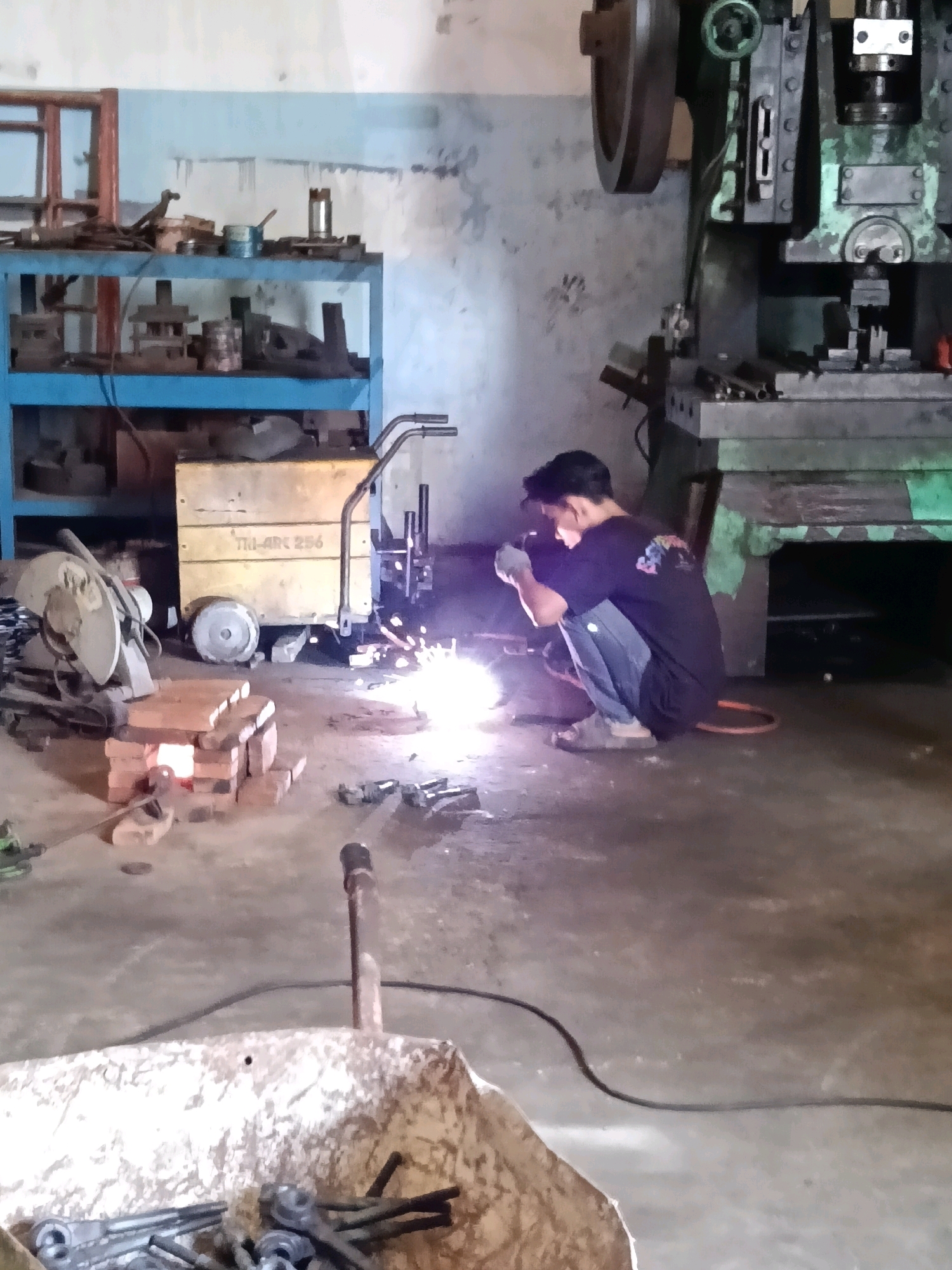 In a dimly lit workshop, likely located in Belimbing, Indonesia, a person is welding. The individual, wearing a dark t-shirt, blue jeans, and light-colored gloves and shoes, is crouched down, focused on the task. Sparks are flying from the welding torch, illuminating the immediate area with a bright glow. The workshop appears to be industrial in nature, with various pieces of machinery and equipment visible. To the right, a large, green metal press machine dominates the background. In the center, a yellow cart with "TRI-ARC 256" visible on its side is positioned near the welding activity. In the foreground, a pile of bricks forms a makeshift support, and to the left, a metal-cutting saw is partially visible. Tools and metal scraps are scattered across the concrete floor, suggesting an active workspace. The walls are rough and textured, painted a pale blue and grey, with visible stains and imperfections.