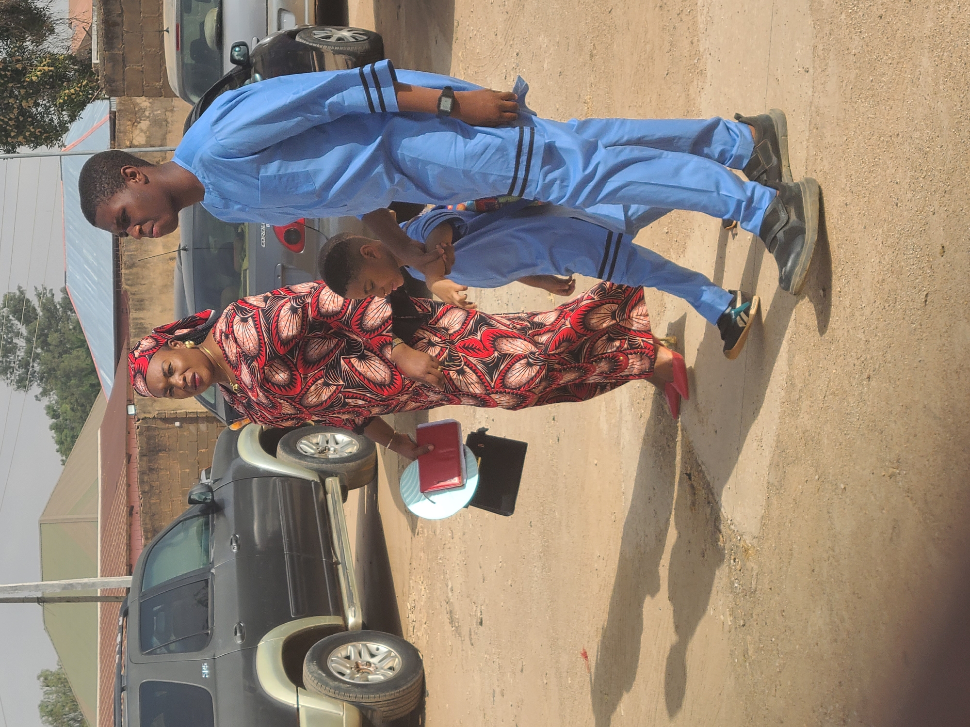 The image shows a woman and two boys standing on a dirt ground in Gura Top, Nigeria. The woman is wearing a red and brown patterned dress with a matching headscarf and red shoes, she is holding a light blue plate with a red book and a black book on it. The boys are wearing light blue outfits with black stripes on the sleeves and trousers. The older boy wears black shoes and a watch. A black SUV is parked behind the woman. In the background, there are buildings and cars.