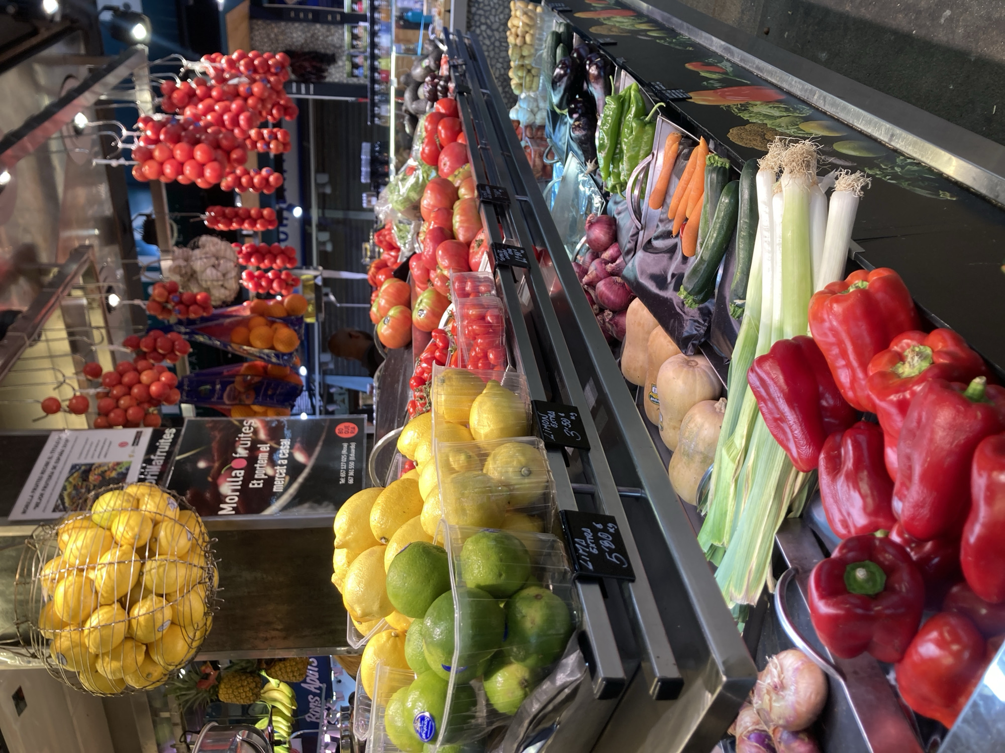 This image depicts a vibrant produce stall at La Boqueria Market in Barcelona, Spain. The stall is overflowing with a colorful array of fresh fruits and vegetables displayed on tiered shelves. Prominently featured in the foreground are bright red bell peppers and a bundle of leeks, with various other vegetables like zucchini and carrots also visible. Higher shelves showcase lemons and limes in plastic containers, while bunches of red cherry tomatoes hang from above. The background reveals more produce, including onions and what appear to be butternut squash, along with other market stalls and customers in the distance. The overall atmosphere is that of a bustling market, rich with the sights and implied smells of fresh, healthy food.