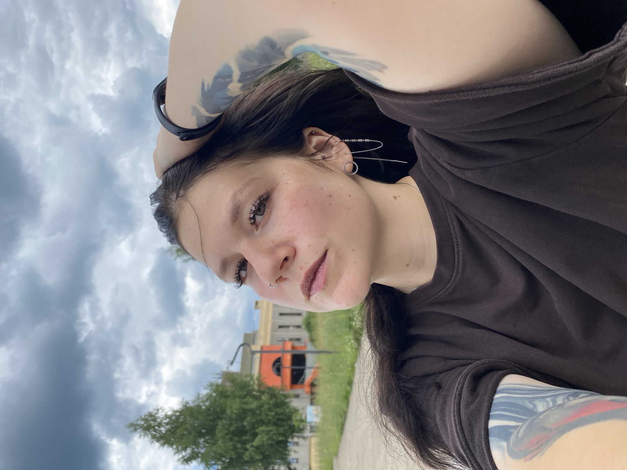 This is a selfie of a young person with dark hair, fair skin, and freckles, looking towards the camera. They are wearing a dark brown t-shirt and have several piercings in their ears and nose. Tattoos are visible on their arms. The person's left arm is bent behind their head, and they are wearing a black fitness tracker on their wrist. The background shows a cloudy sky, a tree, and a building with orange accents, suggesting an outdoor setting. The lighting indicates it is daytime, with overcast weather. The image is captured at an angle, making the orientation appear sideways. There is no visible text in the image. The setting appears to be urban or suburban, possibly in Markovsky, Russia, given the provided location context. The person's expression is neutral, with a slight tilt of the head.