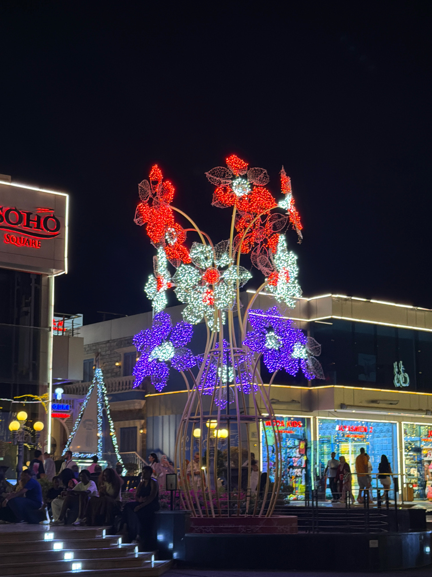 The image depicts a night scene at Soho Square in Sharm El Sheikh, Egypt. A large, illuminated sculpture of flowers made of red, white, and purple lights stands in the foreground. To the left, the illuminated sign "SOHO SQUARE" is visible on a building. In the background, shops and other illuminated structures line the square. Several people are seated on steps in front of the sculpture and walking through the square. The overall atmosphere is festive and vibrant, typical of a popular tourist and entertainment destination at night.