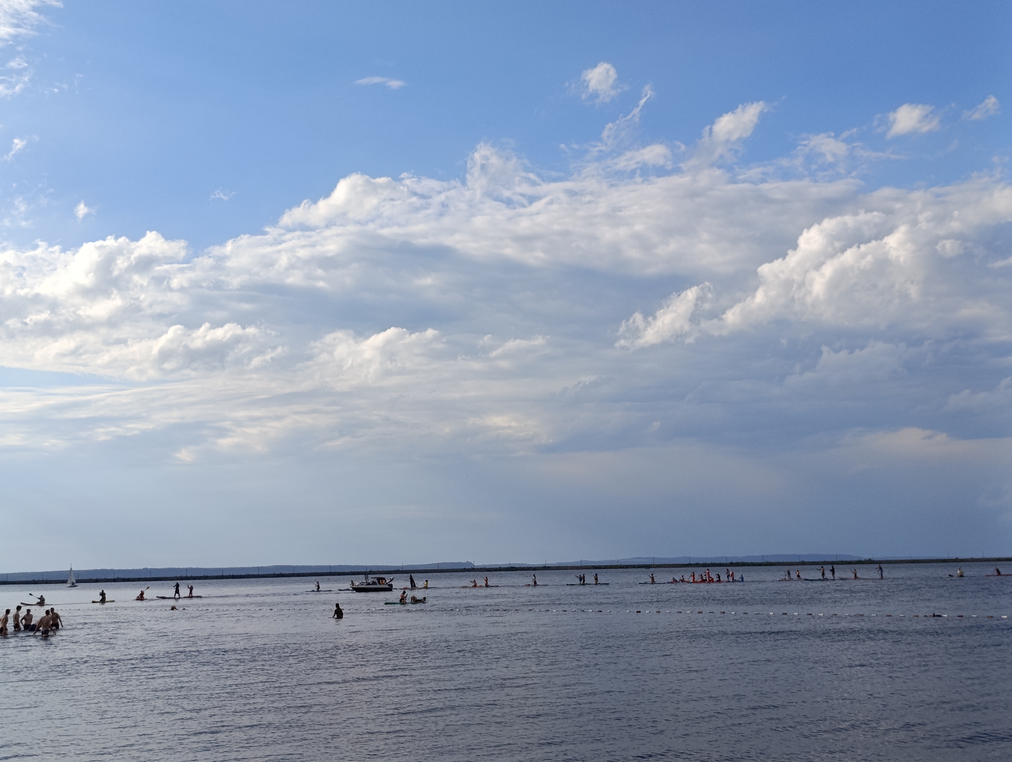 An outdoor, eye-level shot captures a body of water with several people engaged in various water activities under a bright, cloudy sky. The water is dark blue and appears calm. In the foreground, on the left, a group of people are standing in the water near the shore. Further out, multiple individuals are on paddleboards and in kayaks, paddling across the water. A small sailboat is visible in the distance on the left, and a motorboat is positioned on the right side of the frame. A long, low structure, possibly a breakwater or a distant shoreline, stretches across the horizon. The sky is a vivid blue with scattered white clouds, some appearing wispy and others more dense and fluffy. The overall scene suggests a recreational day at a lake or the sea.
