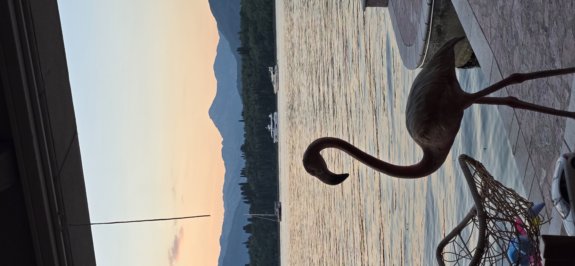 The image depicts a sunset scene at Bogišići, Montenegro. A metal flamingo statue stands on a stone surface overlooking a body of water, likely the Bay of Kotor. The water reflects the sunset colors. In the background, mountains are silhouetted against the sky. Several boats are visible on the water. A net-like object and part of a white object are located on the side.