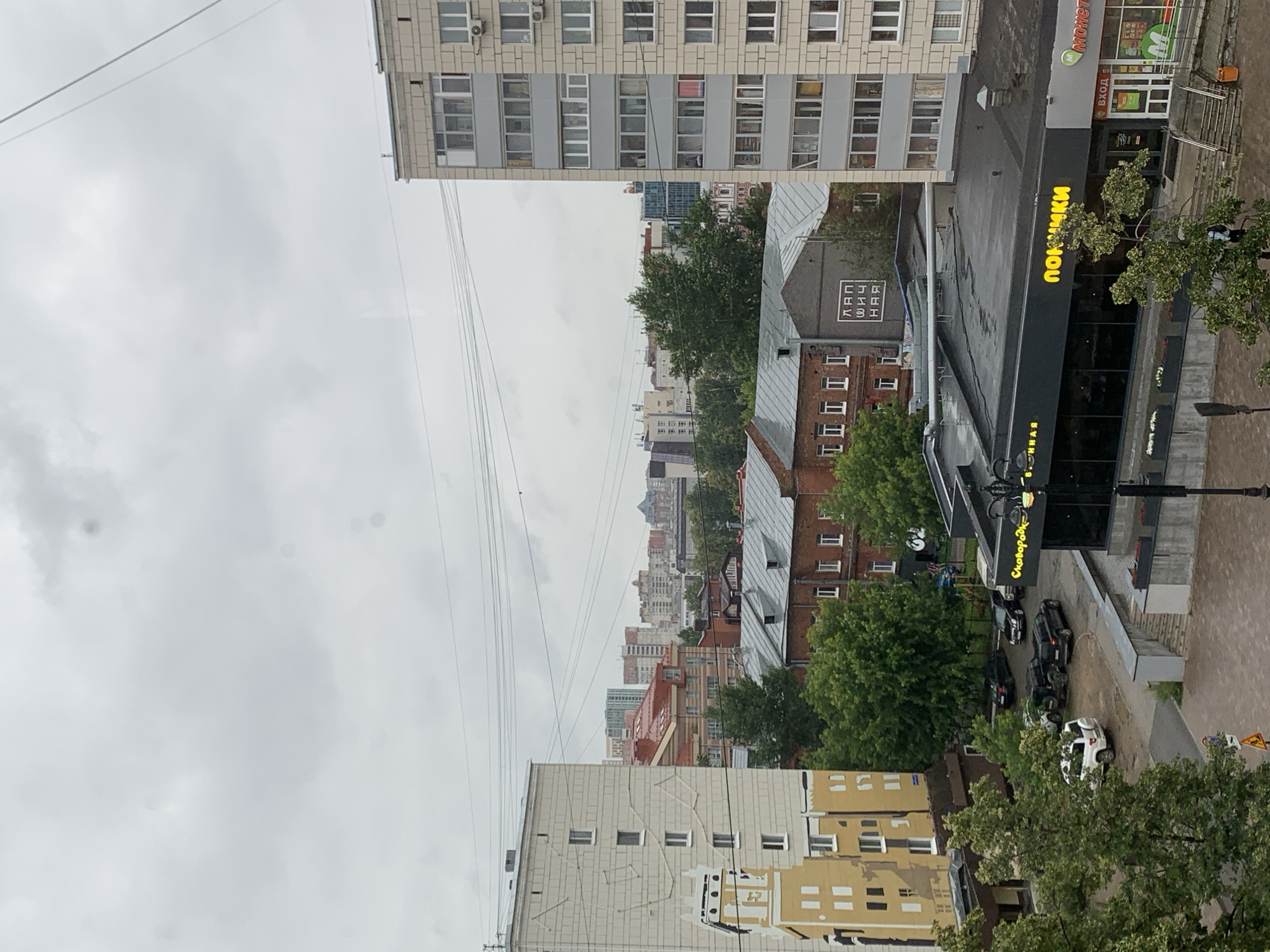 This is a high-angle, slightly tilted shot of an urban street scene in Perm, Russia. The weather appears overcast and possibly rainy, with a muted sky and wet surfaces visible on the street. The image is dominated by buildings of various architectural styles and heights. In the foreground, a modern, multi-story building with a grey facade and numerous windows is prominent. To its right, a lower, darker building with a sloped roof features a prominent yellow sign with Cyrillic text that reads "ПОНИМКИ" (PONIMKI), suggesting it might be a food establishment or store. Another sign above it, partially visible, says "монет" (monet).
Further into the background, a cluster of buildings creates a dense cityscape. These include brick buildings with red roofs and more modern structures with glass facades. Trees with lush green foliage are interspersed throughout the scene, adding a touch of nature to the urban environment.
Several cars are parked along the street, including black SUVs and a white hatchback. The presence of cars and the urban architecture indicate a typical city setting. Utility lines are visible crisscrossing the sky. No people are clearly discernible in the image. The overall mood is subdued, likely due to the cloudy weather and the grayscale tones of the buildings. The visible text primarily relates to business names.
