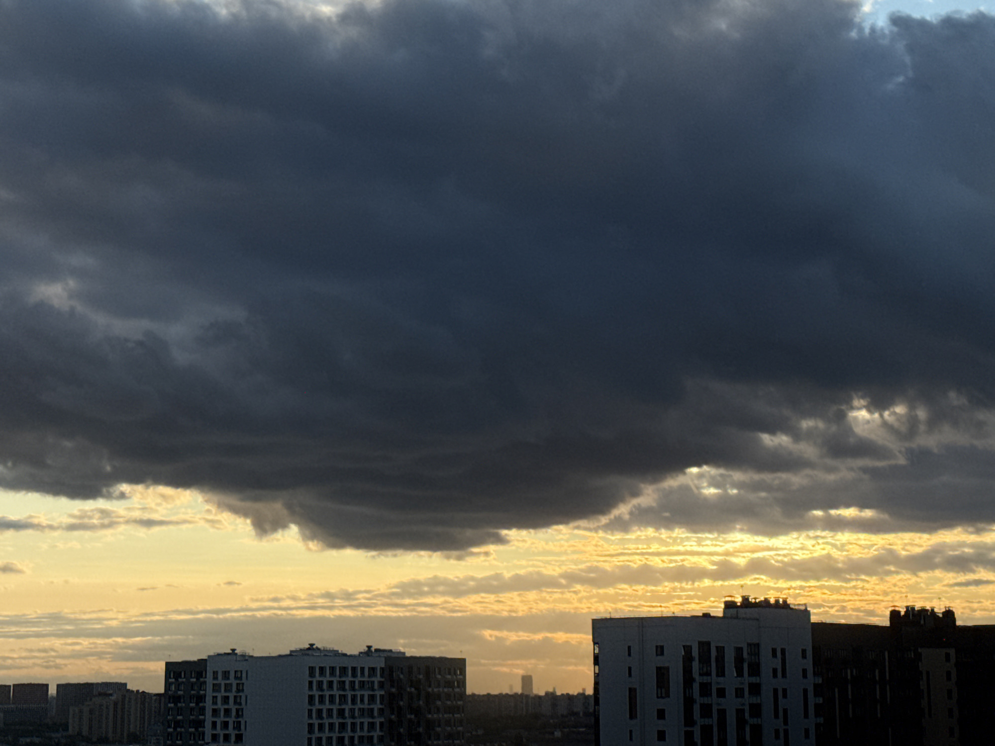 The image depicts an urban skyline under a dramatic sky, likely in Lyubertsy, Russia. Dominating the upper portion of the frame is a vast expanse of dark, ominous clouds, creating a sense of impending weather. Below these clouds, a strip of bright yellow and orange light suggests the sun is setting or has just risen, illuminating a layer of thinner, wispy clouds. In the foreground, the silhouettes of modern apartment buildings are visible, their geometric shapes contrasting with the organic forms of the clouds. The buildings are multi-story structures with numerous windows. The overall impression is one of an urban landscape preparing for or recovering from a storm, with the vibrant sunset hues offering a touch of visual drama against the brooding sky. No people are visible in the image.
