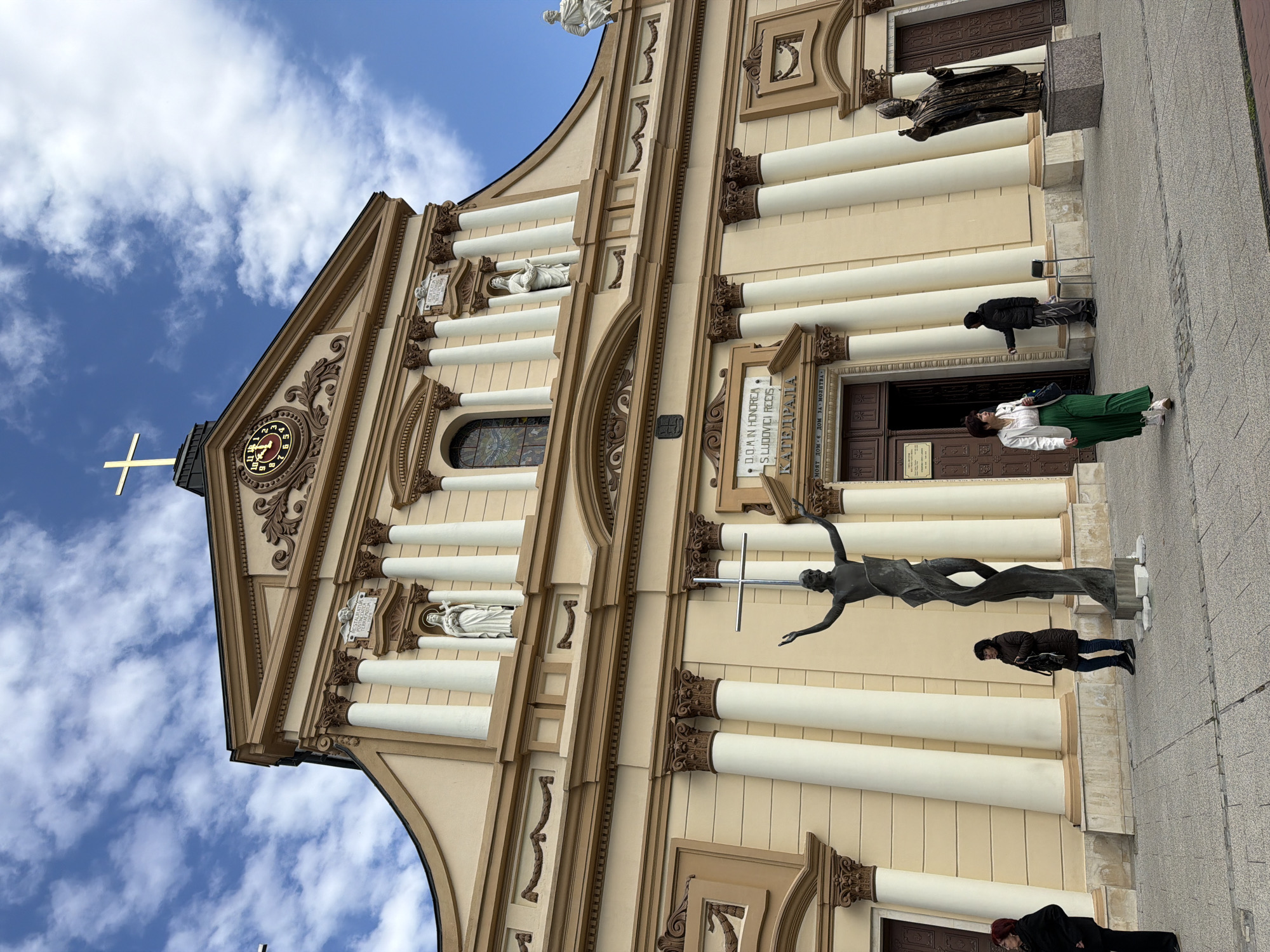 The image depicts the facade of the Cathedral of St. Louis in Plovdiv, Bulgaria. The cathedral is a grand, multi-story building with a beige and brown color scheme. It features numerous white pillars, statues in niches, and ornate detailing around the windows and roofline. A large cross is visible atop the roof.
Several people are visible in the foreground near the entrance of the cathedral. They are standing and appearing to be tourists or visitors. One woman stands out wearing a green skirt. A sculpture is also prominent in front of the cathedral, seemingly depicting a figure in motion with outstretched arms holding a cross. The foreground is a paved area, likely a sidewalk or plaza in front of the building. The sky is visible in the background, featuring blue patches with white clouds.
