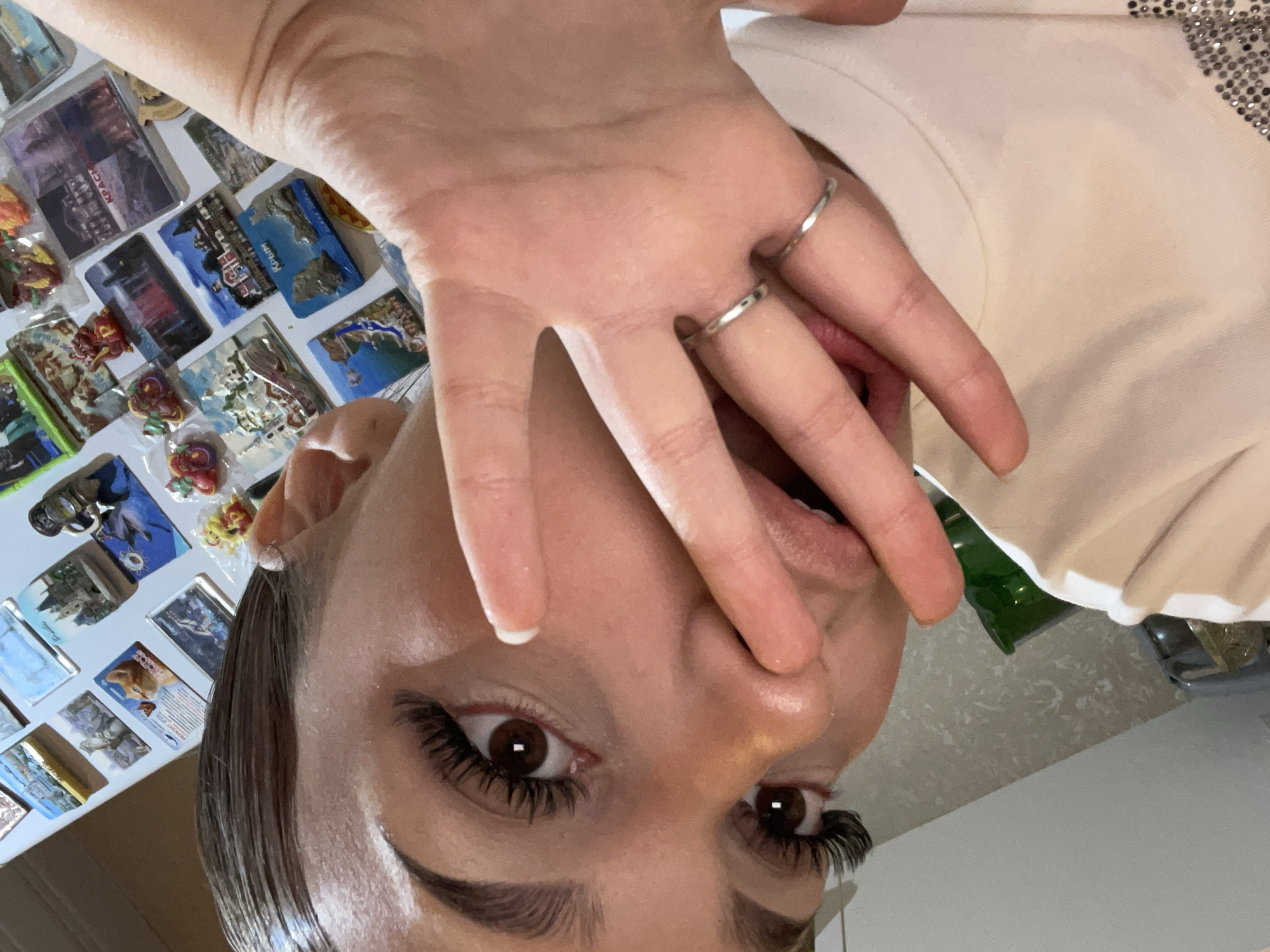 The image is a close-up, high-angle selfie of a person, likely female, with dark hair, brown eyes, and prominent long eyelashes with winged eyeliner. Her left hand is partially covering her mouth and nose, with fingers extended across the lower part of her face. Two simple silver rings are visible on one of her fingers. Her mouth is slightly open, revealing a glimpse of her tongue. She is wearing a light-colored, possibly beige or off-white, top with sparkly, sequined embellishments visible on the upper right side of the frame near her shoulder.
The background primarily consists of a collection of numerous refrigerator magnets displayed on a white surface, likely a refrigerator door. These magnets depict various scenes, including landscapes, buildings, and abstract designs. Visible text on one blue magnet clearly reads "Крым" (Crimea in Russian). Another magnet shows text that appears to include "Красиво" (Beautiful in Russian). The assortment of magnets suggests a personal collection, possibly souvenirs.
Below the magnet display, a sliver of a green object, possibly a bottle, and a neutral-toned surface are visible. The image's perspective and the subject's gaze suggest a direct and possibly playful engagement with the camera. Given the visible text "Крым" and the provided location context, the scene is likely set in Russia, with the magnets referencing Crimea. The time of day and weather are not discernible, but the setting appears to be indoors.