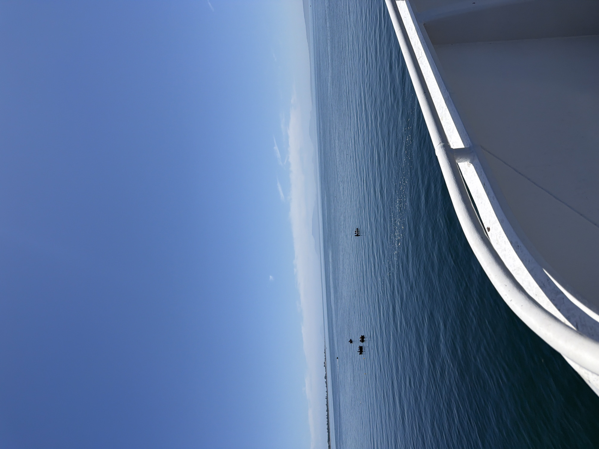 The image shows a wide expanse of calm blue water under a clear blue sky. In the distance, a hazy shoreline with some greenery is visible, possibly indicating land or islands. Several small boats are scattered across the water, some with people in them, suggesting recreational or fishing activities. In the foreground, the edge of a white structure, likely the railing of a boat or ferry, curves into the frame. The overall scene depicts a serene day on the water. While the image does not provide explicit details to pinpoint the location within Delta, Canada, the presence of calm, open water and a distant shoreline is consistent with coastal or bay areas in the region.