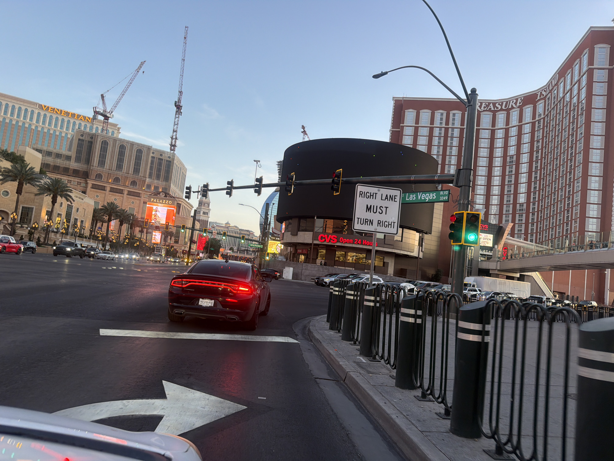 The image captures an urban scene in Paradise, Nevada, United States. The time of day appears to be early evening, as indicated by the sky's color. A black sedan, with license plate "NZ-230-878" is in the foreground, positioned on a street marked with a right-turn arrow. The car's brake lights are illuminated.
Visible structures include the Venetian and Treasure Island hotels, both iconic landmarks in Paradise. Construction cranes are near the Venetian, suggesting ongoing construction or maintenance. Street signs indicate "Las Vegas Blvd" and the street number "3249". A large electronic sign, partially obscuring the view, is present, along with a "Right Lane Must Turn Right" sign. A CVS store is also visible with the text "CVS Open 24 Hours."
Traffic lights are present, showing green on the right side and red on the other side of the intersection. A line of bollards is present near the sidewalk. Cars are parked along the street in the middle ground. The overall environment suggests a bustling urban intersection in a tourist area.