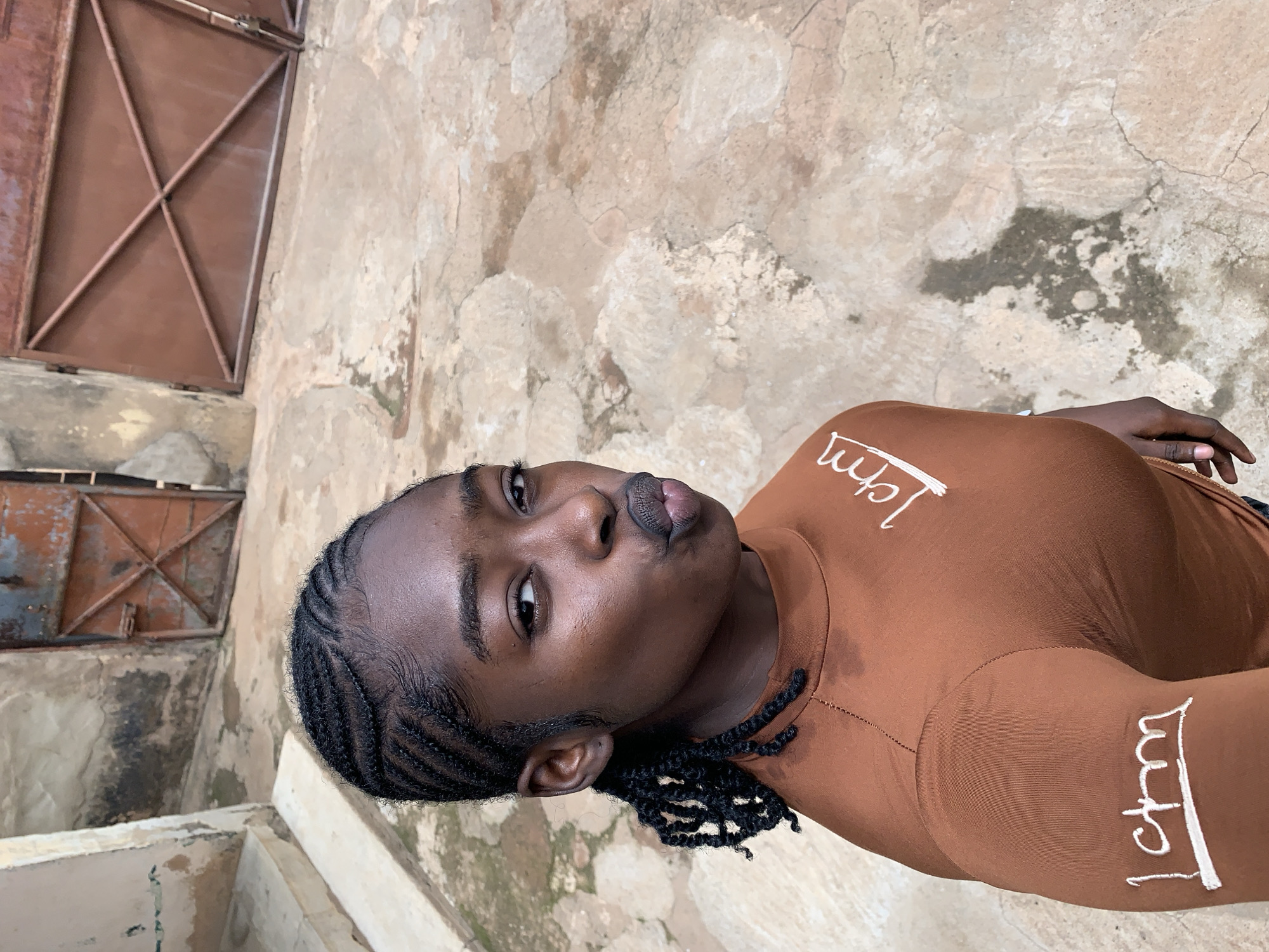 A young Black woman is featured in a close-up, high-angle selfie taken in Rafin Par, Nigeria. She directly faces the camera with pouted lips, her head slightly tilted. Her dark brown skin has a subtle sheen, and her hair is styled in cornrows pulled back from her forehead, transitioning into twisted braids gathered behind her left ear. She wears a long-sleeved, mock-neck brown top with stylized white "1cm" text on both her right sleeve and upper chest. The outdoor setting includes a rough, light brown ground surface exhibiting cracks and dark, discolored patches. In the background, a light-colored, weathered concrete wall with a staircase is visible on the left. Further back are two rusty brown metal gates: a single, partially open gate with a diagonal crossbar on the left, and a larger, closed double gate featuring an "X"-shaped support structure on the right.