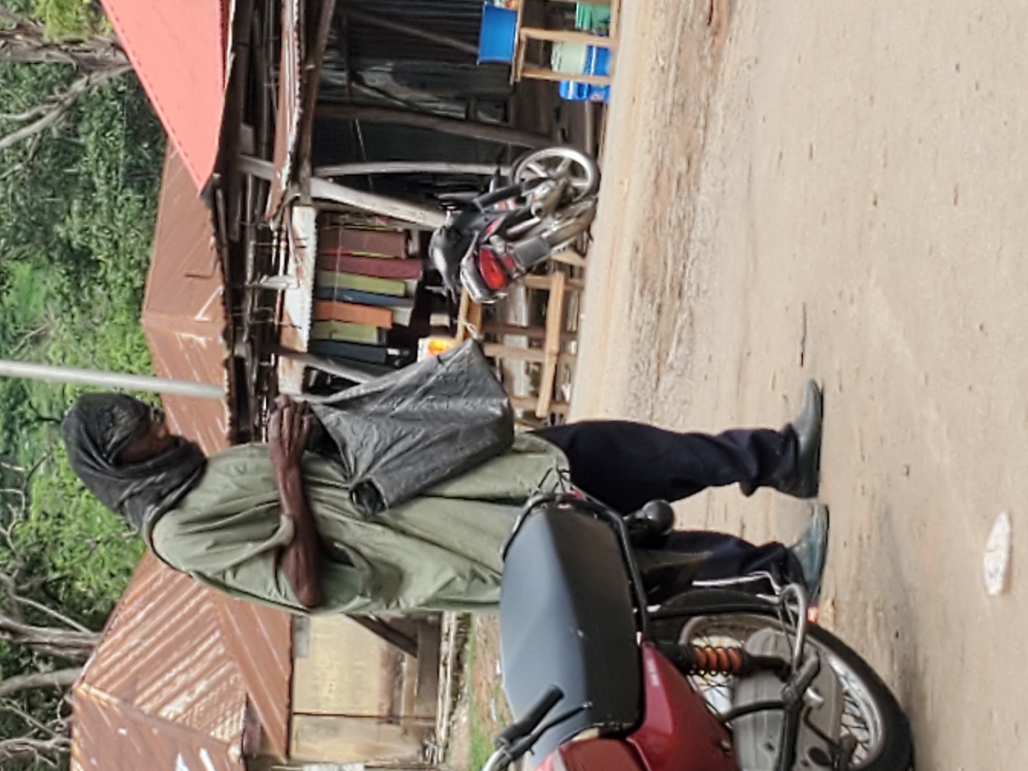 The image depicts a man standing next to a motorcycle in Mararaba, Nigeria. The man is wearing dark pants and black shoes, a light green shirt, and a dark head covering. He has a black plastic bag slung over his shoulder. He is standing on a dirt road next to the motorcycle, which is primarily red and black. Behind the man and the motorcycle is a simple structure, possibly a shop or home, with a rusted corrugated metal roof. Various items, including colorful fabrics and plastic containers, are visible within the structure. Another motorcycle is parked inside the structure. Trees are visible in the background. The road appears to be unpaved and dusty.
