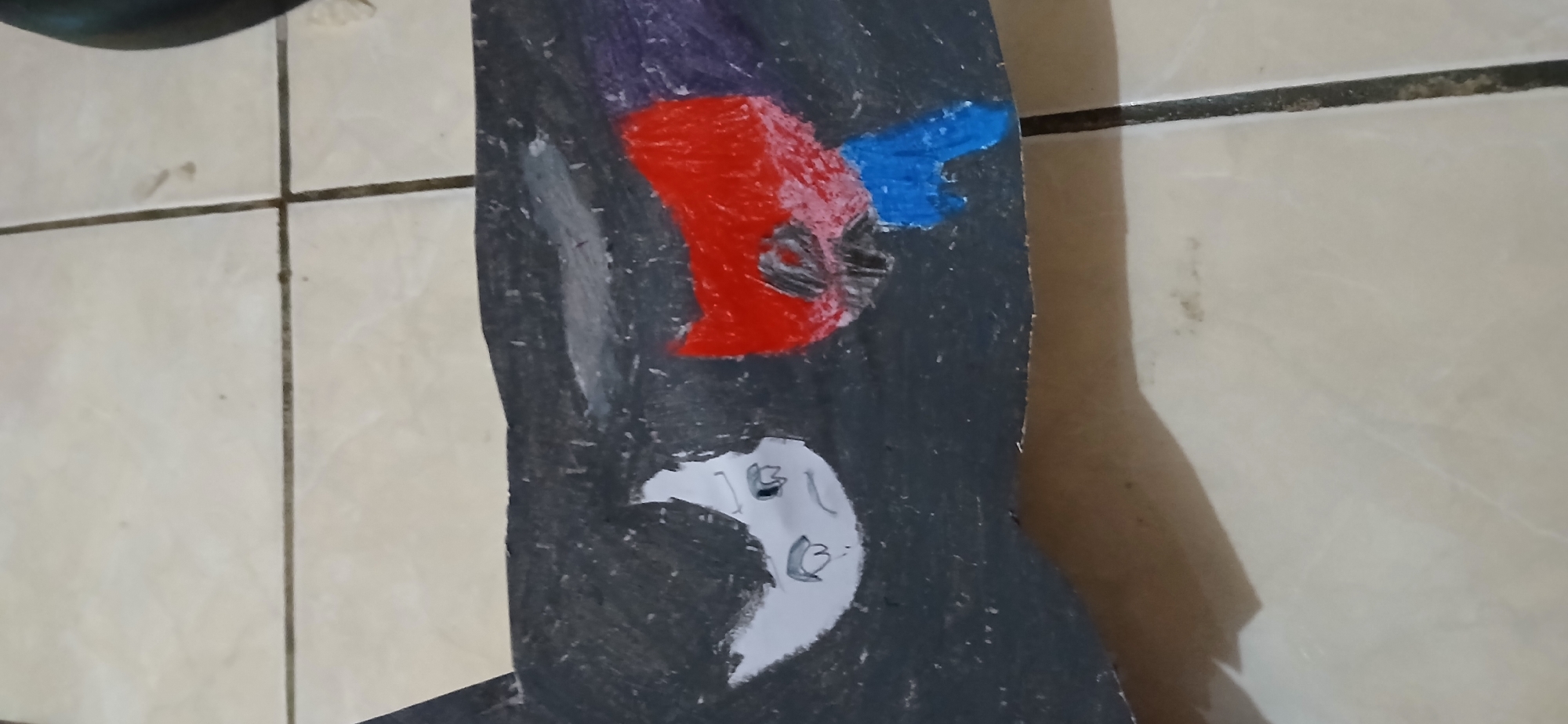 The image shows a piece of dark, possibly cardboard, material with drawings on it. On the left is a drawing of a white face with black hair and a sad expression, with large grey eyes and a small mouth. To the right of the white face is a drawing of a red, bat-like mask with a black outline and a blue shape underneath, which could be part of a costume or character. The drawings appear to be made with colored pencils or crayons. The material is lying on a tiled floor, which is light beige with dark grout lines. The background shows more of the tiled floor and the edge of a dark object on the far right. The scene is indoors. There are no visible landmarks or indicators that would allow for localization to Samarinda, Indonesia.