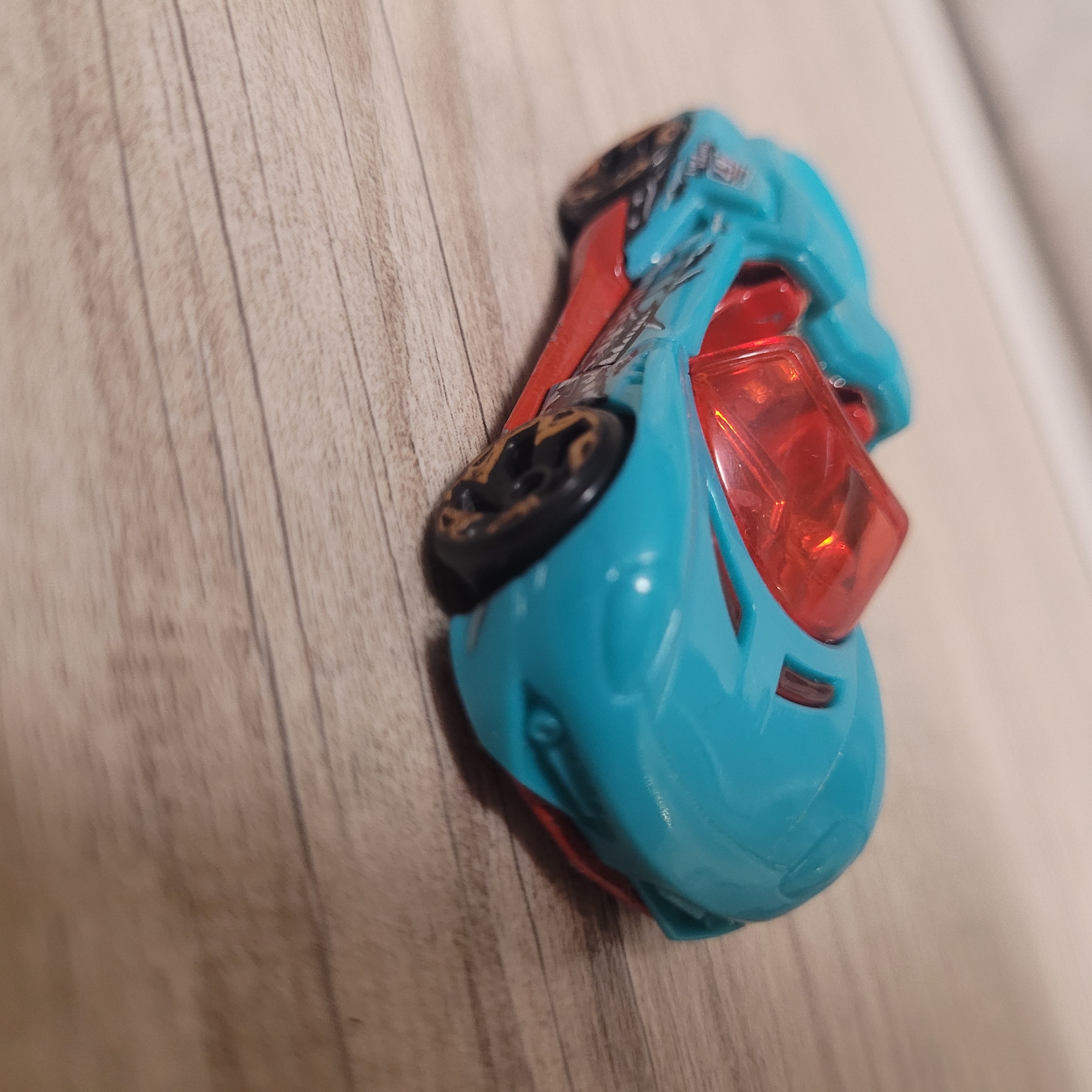 A turquoise and red toy car is shown on a wooden surface. The car is a low-profile sports car with a red transparent windshield and interior. It has black wheels with gold accents. The surface appears to be a tabletop or floor with a wood grain pattern. The image is a close-up shot, with the car occupying the majority of the frame. The lighting is soft and appears to be coming from the side, casting subtle shadows.