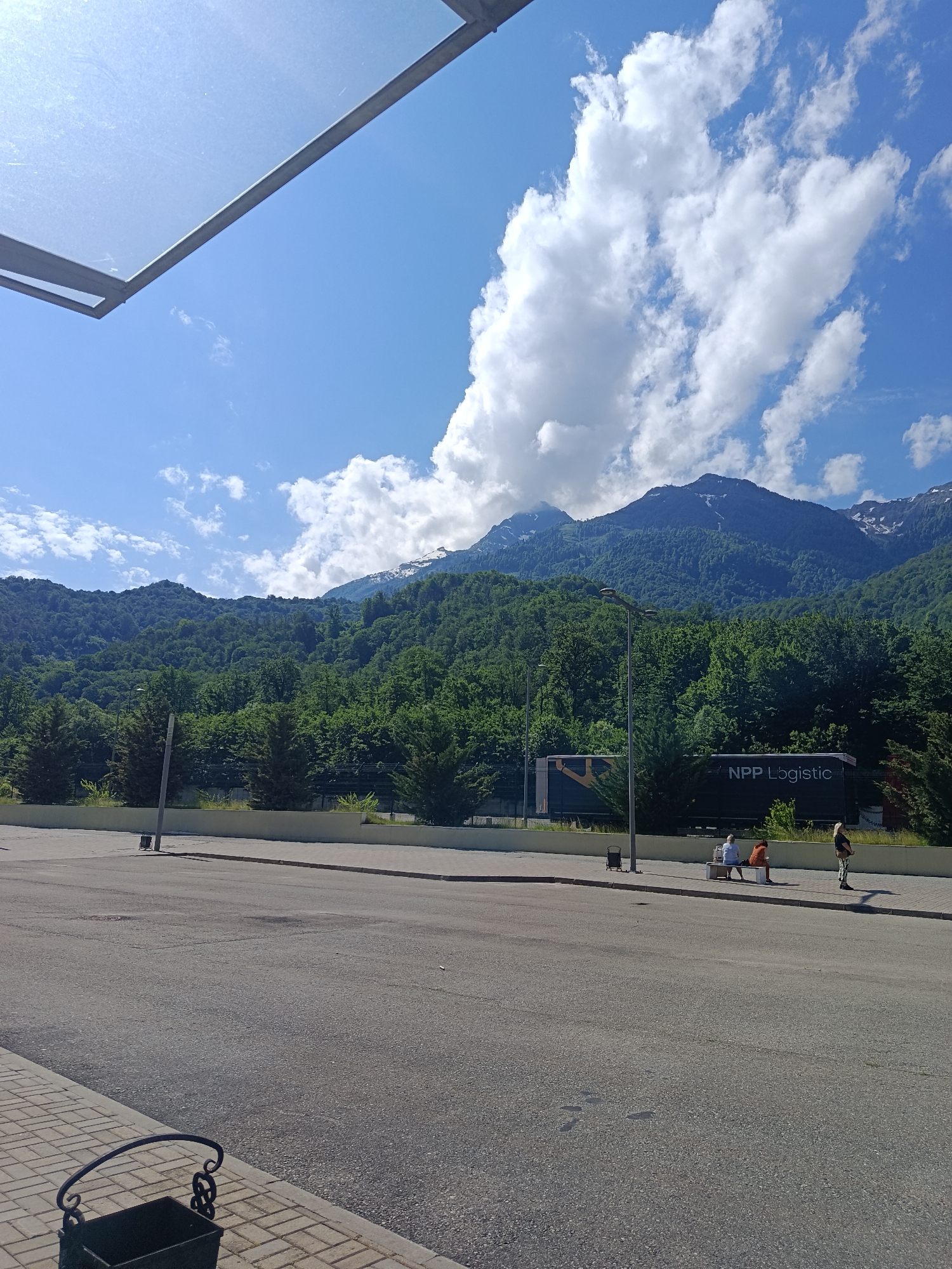 This is a medium shot of a paved area with a backdrop of lush green mountains under a bright blue sky with scattered clouds. In the foreground, a dark asphalt road is visible, with a pedestrian walkway made of light-colored interlocking pavers to the left. A dark metal trash receptacle with decorative scrollwork sits on the paver walkway. Further back, a light-colored retaining wall separates the paved area from a zone with sparse greenery and trees. Beyond the wall, a dark truck with "NPP Logistic" written on its side is parked. Three individuals are visible near the truck, with two seated on a bench and one standing. The mountain range in the background is densely covered in trees, with some patches of snow on the higher peaks. The scene appears to be a public space in Estosadok, Russia, likely a transit or resting area with a view of the natural landscape.