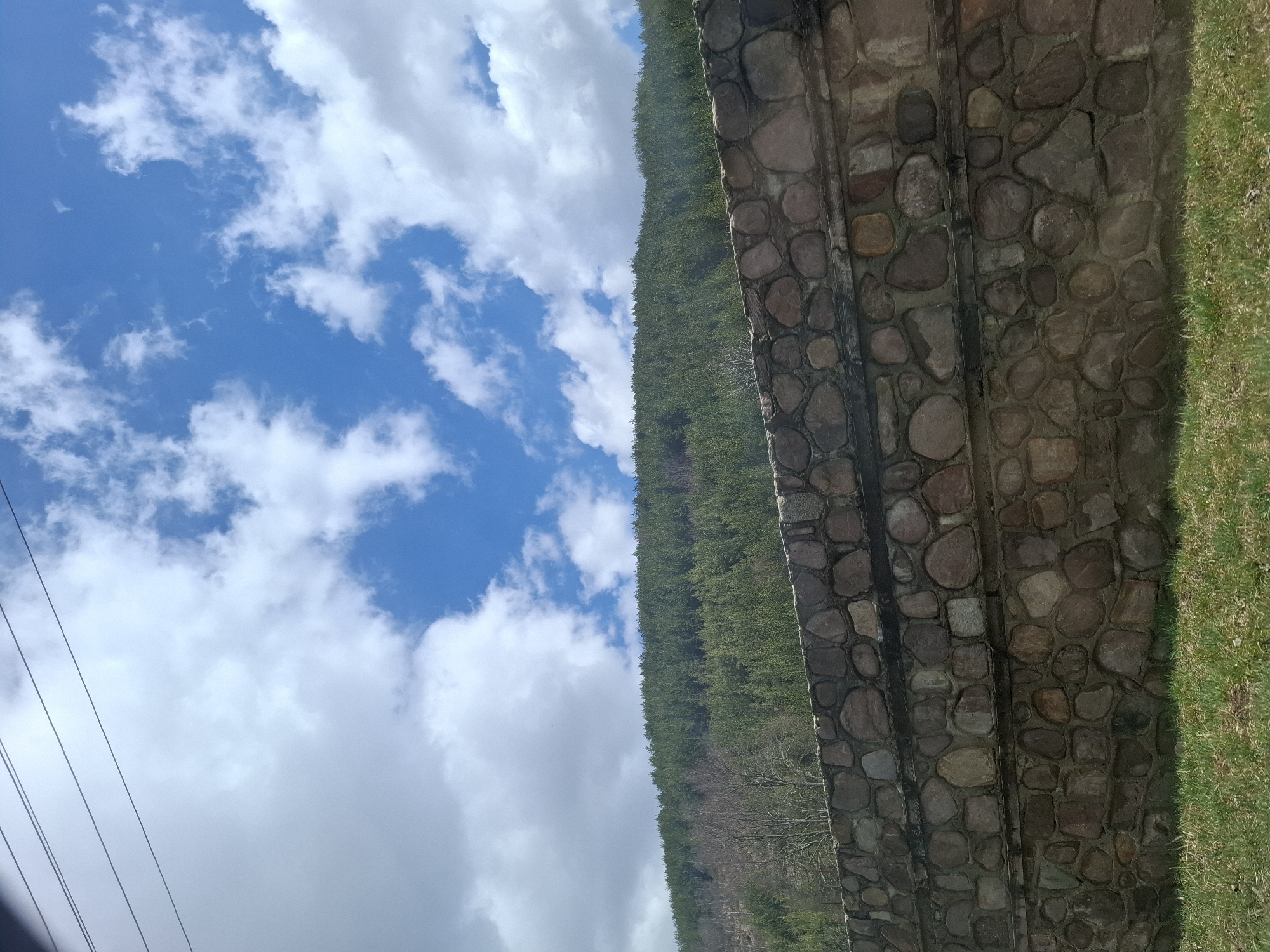 The image shows a landscape in Gintsi, Bulgaria. A stone wall runs vertically on the right side, next to a patch of green grass. A densely forested hill slopes upwards to the left of the wall. The left side of the image shows a blue sky dotted with white clouds. There are also power lines in the corner of the image.