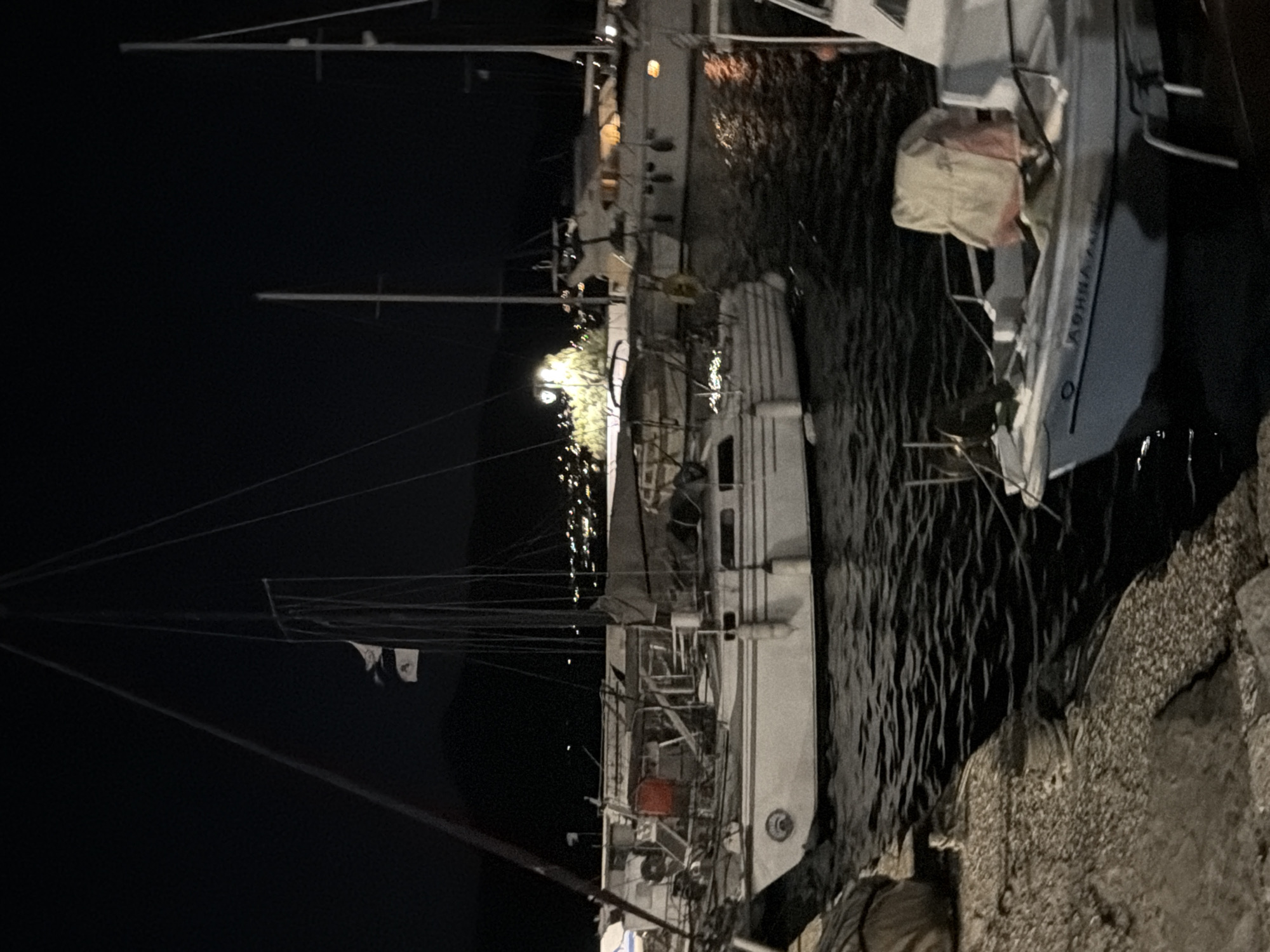 This is a nighttime photograph of a marina in the Municipal Unit of Palairos, Greece. Several sailboats are docked, their masts silhouetted against a dark sky. The foreground features a stone pier. In the middle ground, a white sailboat with a red accent is prominently displayed. Further back, another, larger white boat is visible, its windows illuminated with warm light. The background shows a faint, distant line of lights, suggesting a town or settlement. The water is dark and slightly rippled, reflecting some of the ambient light. The overall impression is one of a peaceful, tranquil harbor at night.