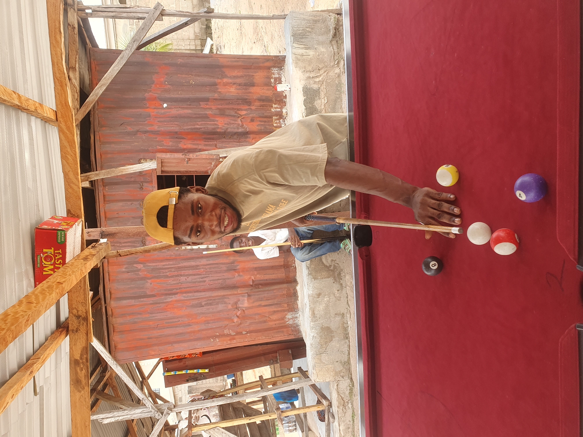 The image depicts two young men engaged in a game of pool in an outdoor setting, likely Lokoja, Nigeria. The foreground is dominated by a red pool table with several balls scattered across its surface, including a black 8-ball, a solid yellow ball, a white ball with a red stripe, a solid red and white ball, and a purple 4-ball. One man, wearing a light brown t-shirt and a yellow baseball cap, is actively playing, holding a cue stick and positioned to strike a ball. He is looking towards the camera with a slight smile. In the background, another man in a white shirt and blue jeans sits on a concrete ledge, holding a cue stick. Behind them, a corrugated metal structure with wooden supports forms a makeshift roof and wall. A red box labeled "TASTE TOM" is visible on the wooden beams above. The environment appears somewhat rustic, with a rough concrete floor and a sandy area in the distance.