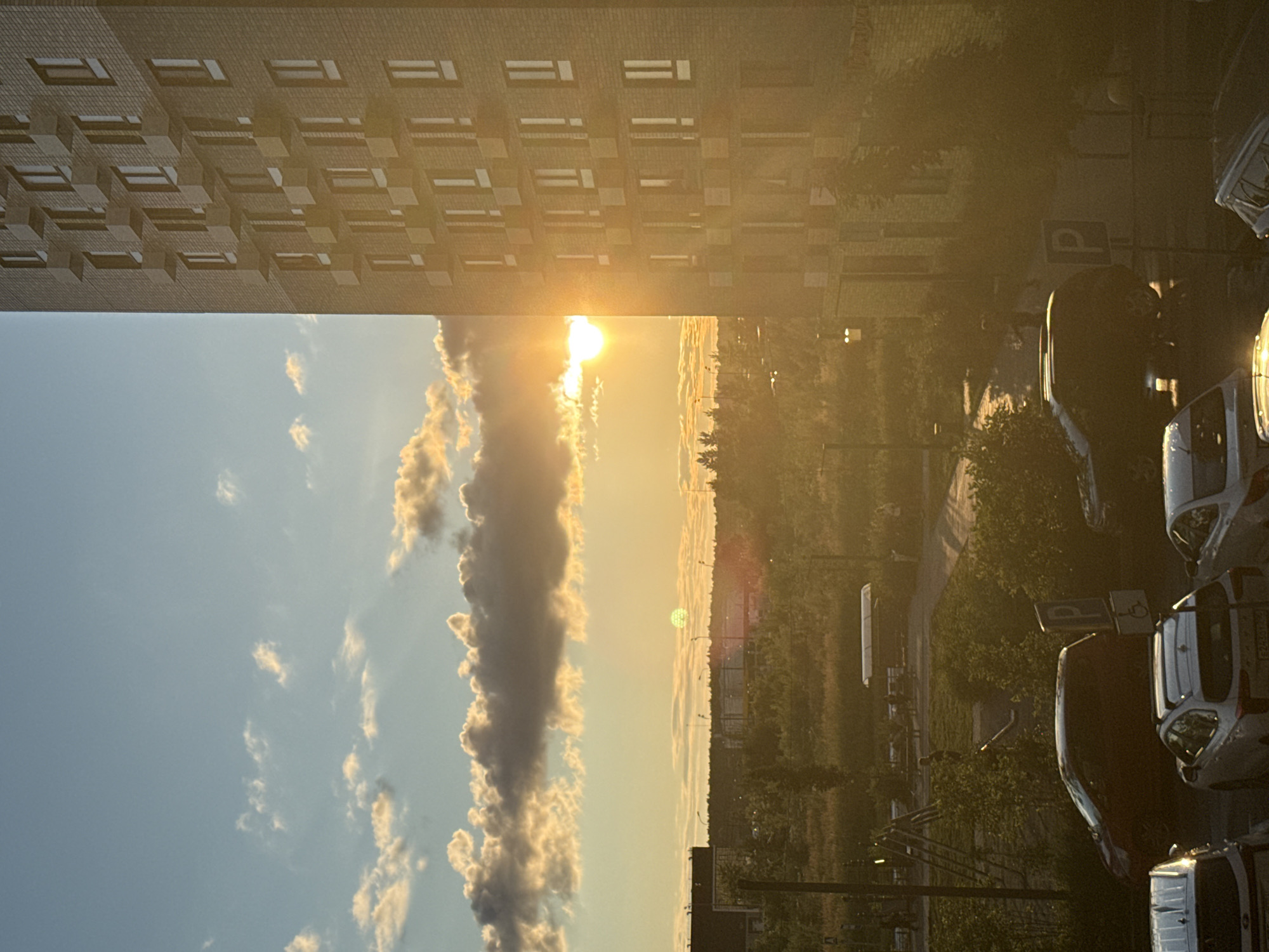 The image depicts a sunset view from an apartment building in Drozhzhino, Russia. The sun is setting behind a treeline and some buildings in the distance, casting a warm glow over the scene. A tall, modern apartment building with visible windows and balconies occupies the right side of the frame. In the foreground, several parked cars are visible, along with a children's playground with a slide and climbing structure. The sky is a mixture of blue with scattered, golden-tinged clouds, illuminated by the setting sun. The overall environment appears to be a residential area with interspersed green spaces.