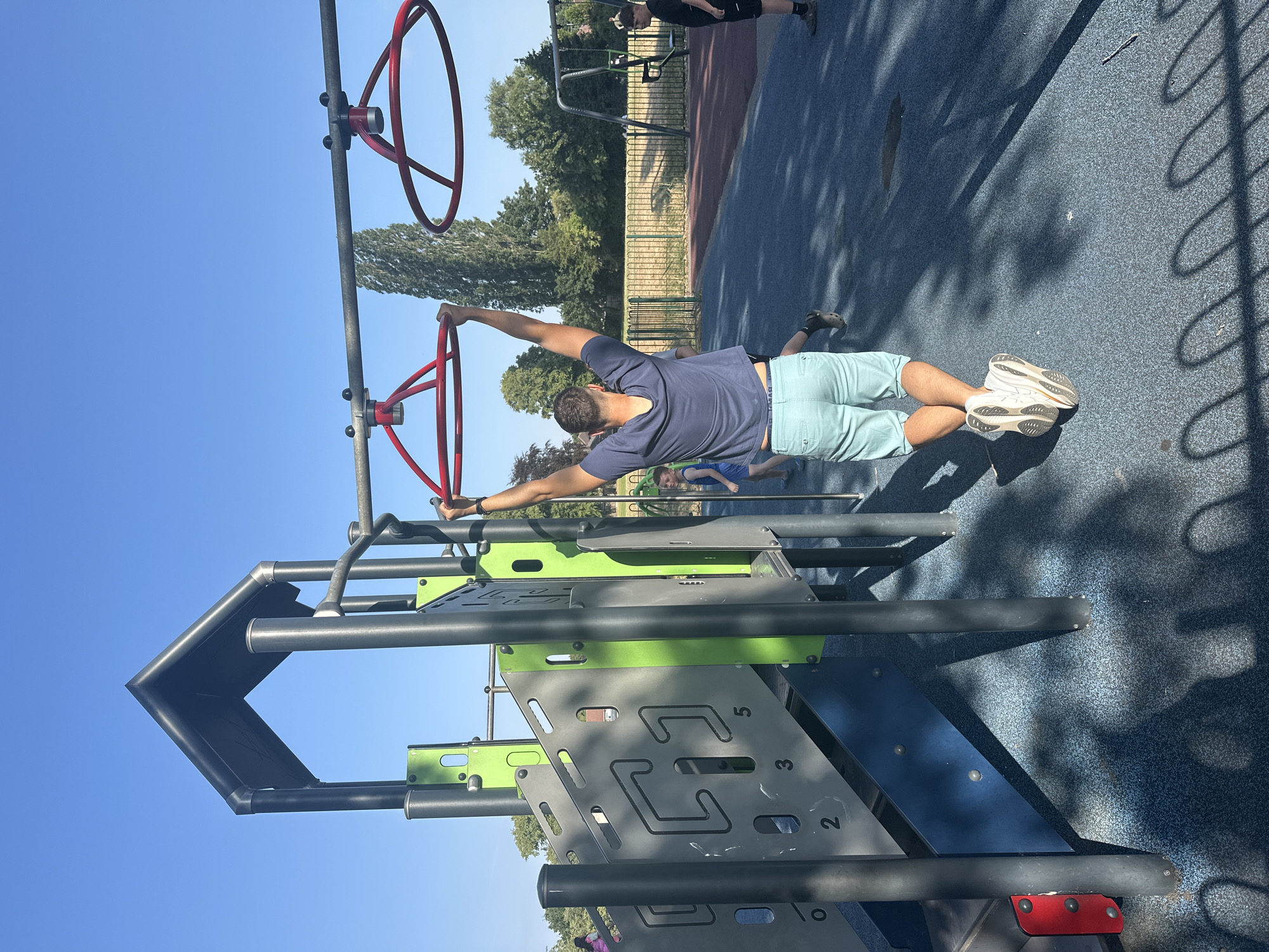 A man is performing pull-ups on a red monkey bar ring in a playground. He is wearing a dark blue t-shirt and light blue shorts. In the background, there is a playground structure with a slide, a swing set, and other children. The playground has a blue rubber surface and is surrounded by a fence. The scene appears to be in a park on a sunny day. The specific location is not discernible from the image alone.