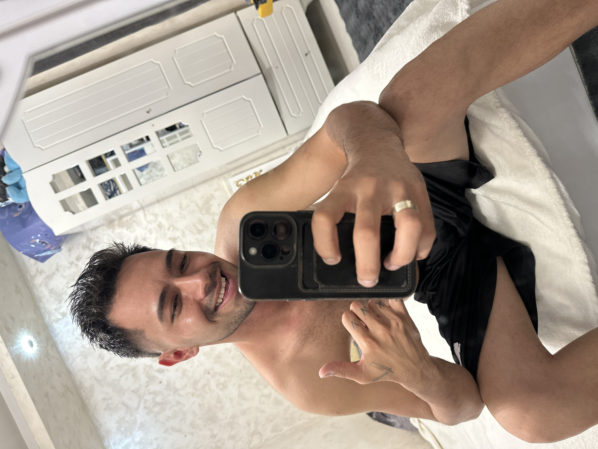 A male individual, appearing to be in his mid-20s to early 30s, is depicted taking a mirror selfie indoors, in a room located in Sofia, Bulgaria. He has short, dark, textured hair with faded sides and a short beard/stubble. He is smiling broadly, exposing his teeth. His torso is bare, and he is wearing black athletic shorts. He holds a black smartphone, likely an iPhone due to the triple camera lens array, in his right hand, angled towards the mirror. A silver-toned ring is visible on the ring finger of his right hand. His left hand is raised, giving a "thumbs up" gesture, and a stylized tattoo of a straight razor is visible on the back of this hand, along with smaller faded tattoos on the fingers.
The environment is a bedroom or personal space. The walls are light-colored with a subtle textured pattern, possibly stucco or wallpaper. A bright, recessed circular spotlight is visible on the ceiling. In the background, to the right of the subject, is a white two-door wardrobe or cabinet. The left door features multiple small glass panes reflecting the room, while the right door is solid with decorative vertical panels. A blue fabric item and other objects are resting on top of the wardrobe. Below the wardrobe doors, two drawers are visible. The subject is seated on a white, soft surface, which appears to be a bed covered with a white blanket or duvet. A dark grey rug or carpet is visible on the floor in the lower right portion of the image.