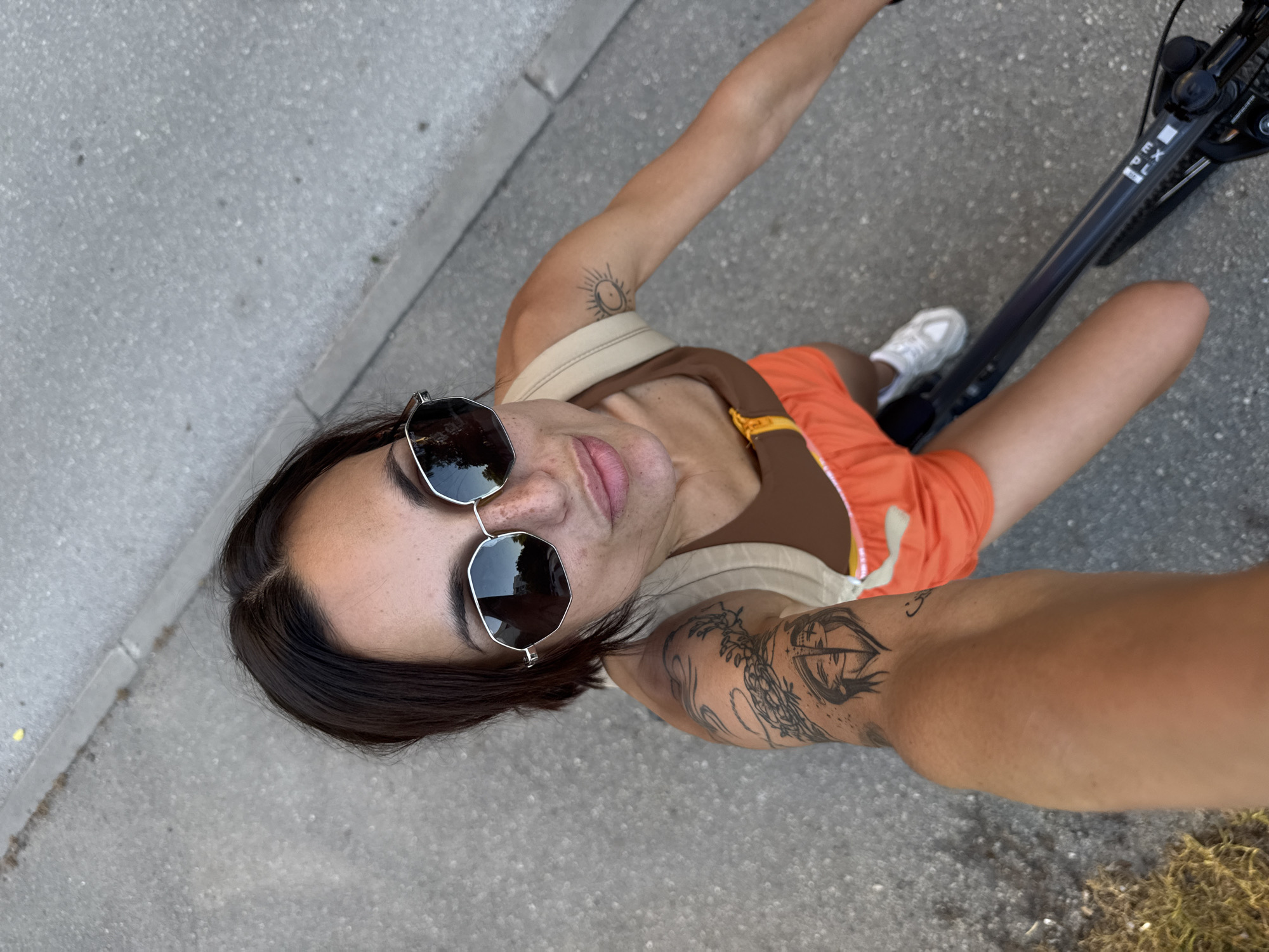 An overhead, close-up, selfie-style shot shows a woman with dark hair and fair skin, wearing sunglasses, a brown top, and orange shorts. She has tattoos on her left arm, including one depicting a split face. She is on a black bicycle, with her right leg bent and her left leg extended. The background is a paved surface, likely a street or sidewalk, with a curb to the left. The scene is outdoors, suggesting a sunny day. There is no specific indication of the location being Pleven, Bulgaria.