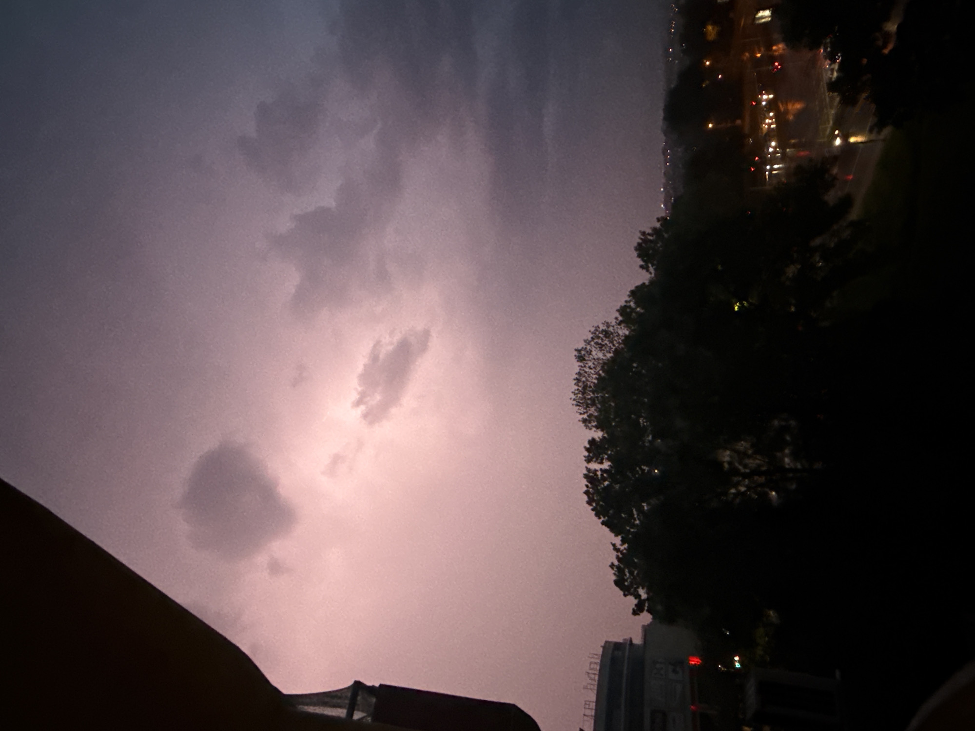 The image presents a nighttime scene with a dramatic sky likely due to a storm. The sky is predominantly purple and grey, with visible clouds. A potential lightning flash is partially visible through the clouds.
On the right side of the image, there's a dark mass suggesting trees or foliage. Further in the distance, a blurred line of city lights is visible, indicating an urban area. These lights appear to be streetlights and building lights.
In the foreground, at the bottom of the image, parts of a building or structure are visible, appearing dark and silhouetted.
Based on the provided information, it is impossible to confirm that the location is Varna, Bulgaria as there are no unique identifiers present.