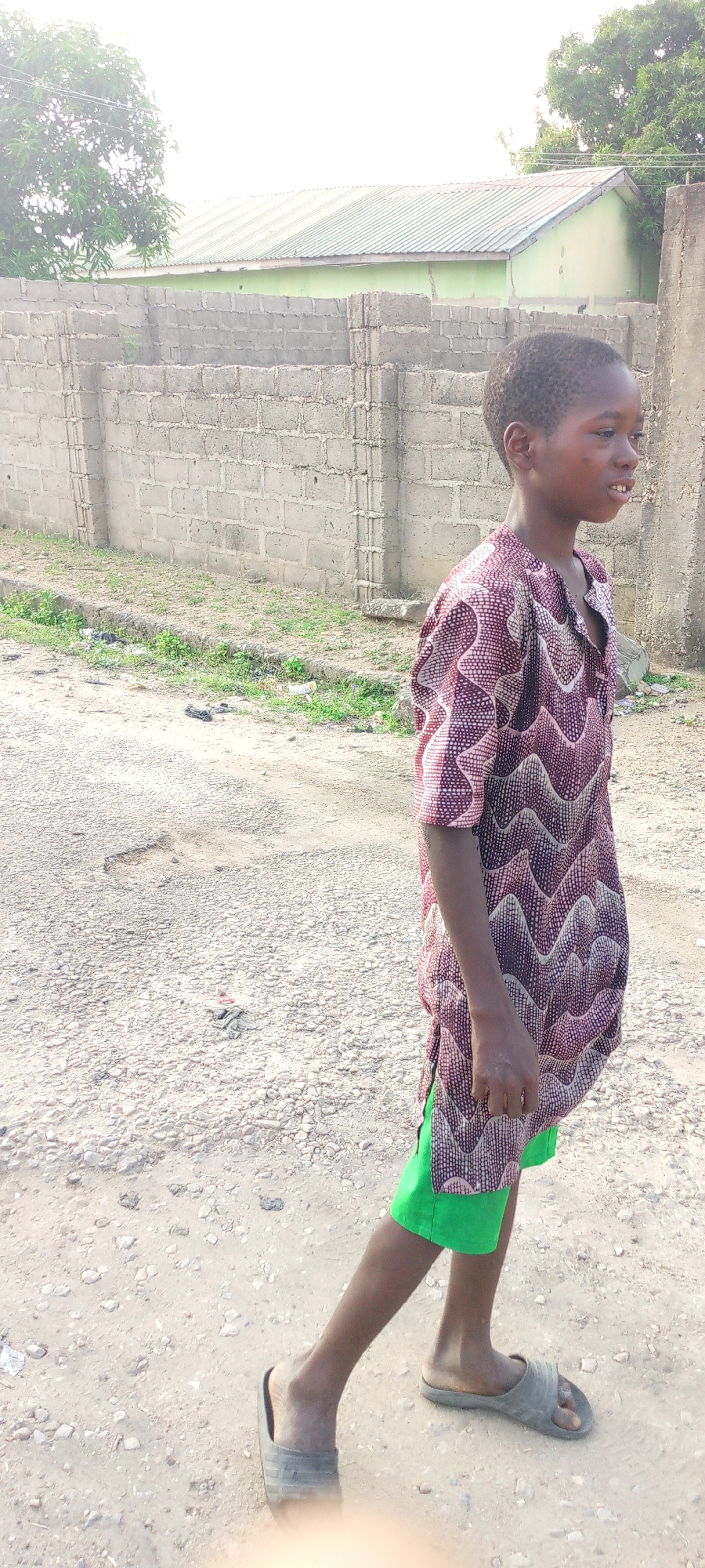 An adolescent boy stands on a dusty street in Adankolo, Nigeria. He is wearing a patterned, maroon and white short-sleeved shirt and green shorts. He is also wearing dark-colored, open-toed sandals. He is facing right, looking off into the distance. The road is unpaved and gravelly. A low brick wall runs along the left side of the road. A building with a green roof is visible behind the wall. Trees are visible beyond the building. A concrete pillar stands at the right edge of the frame.