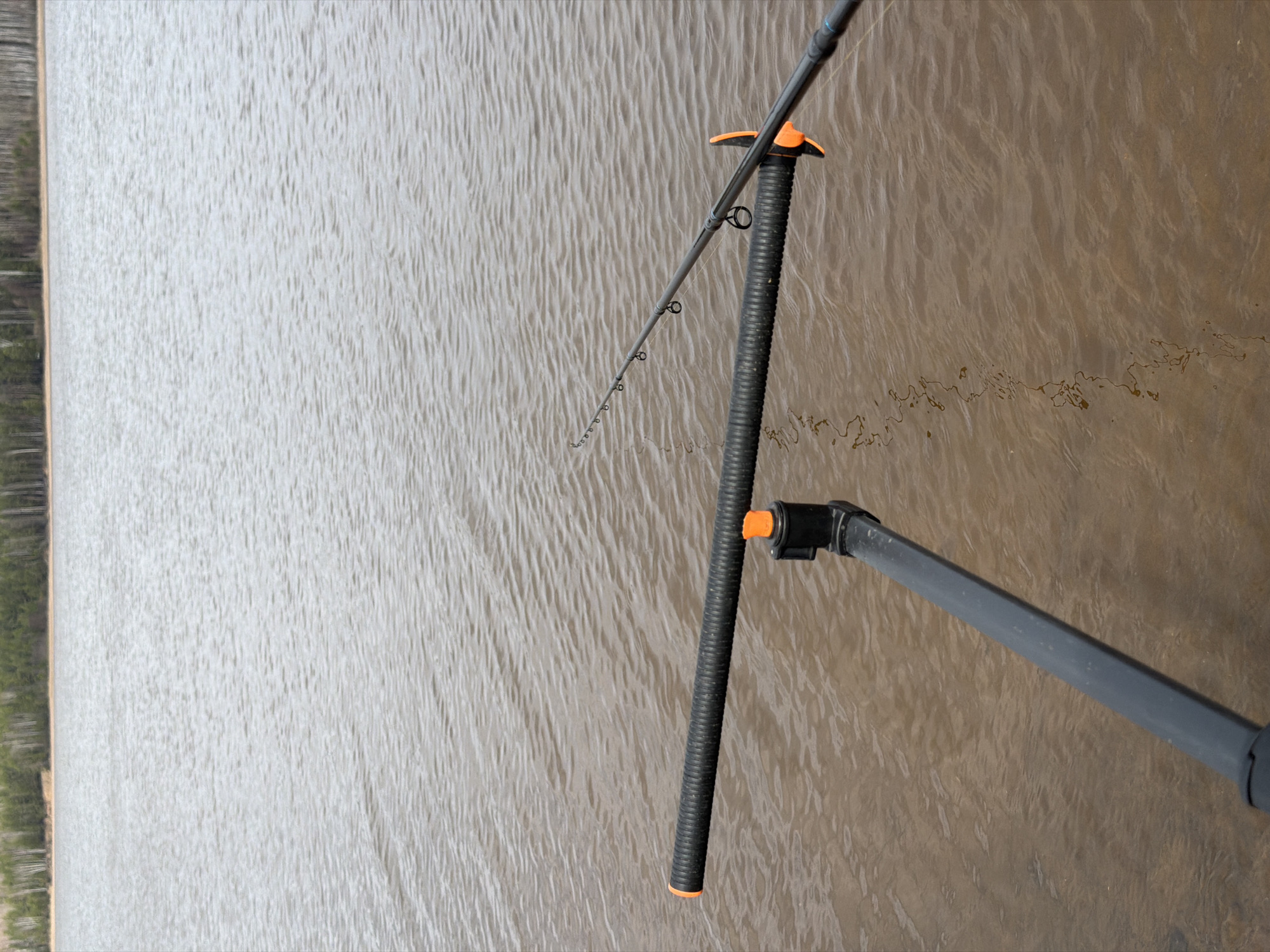 The image depicts a fishing rod in a holder, positioned near a body of water. The rod is thin and gray, with several guides along its length. The holder is black and orange, seemingly made of ribbed material. The water is brown and slightly rippled, suggesting a light breeze. The background shows a blurred shoreline with trees. The location is likely near a body of water, but the specific location in Chaykovsky, Russia, cannot be determined from the image alone. There are no people visible in the image.