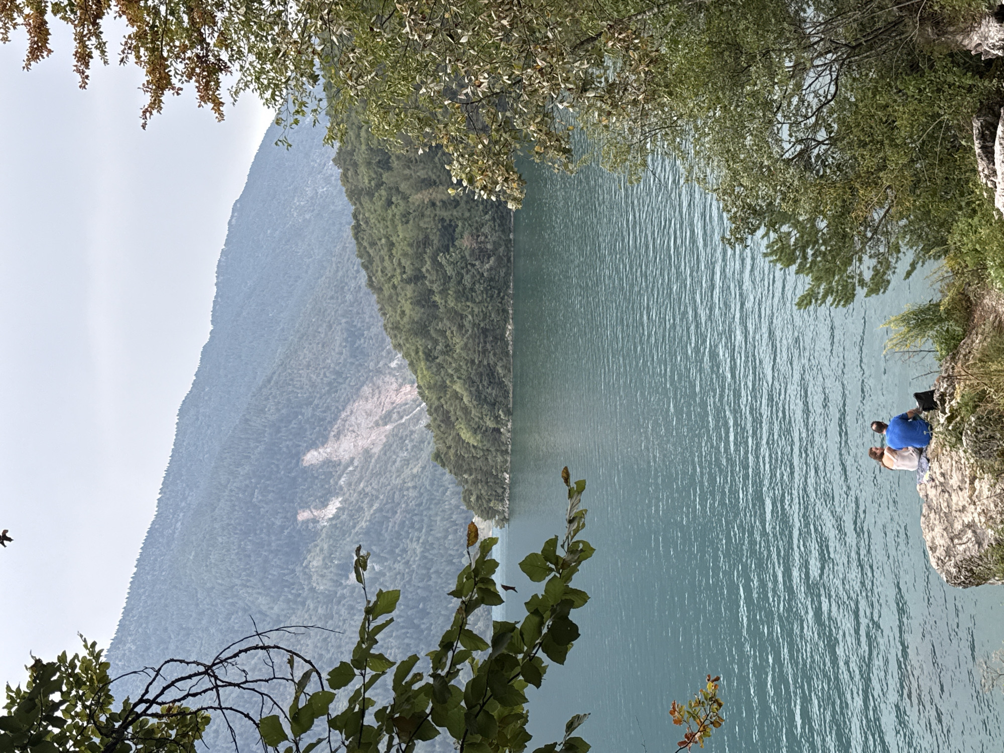 An aerial shot captures a tranquil scene by a large, vibrant blue lake, framed by lush green trees on either side. In the distance, majestic mountains rise, their slopes covered in dense forests. A large portion of the mountain on the far side is bare, revealing a pale, rocky scar.
In the foreground, a couple sits on a rock at the edge of the lake, their backs to the camera. The woman wears a white dress, and the man sports a blue t-shirt. They appear to be engaged in conversation or simply enjoying the view. A small pile of belongings, including a black bag, rests beside them on the rock.
The water's surface is rippled, reflecting the soft, overcast sky. A lone bird is visible in the upper portion of the frame, soaring against the pale sky. The overall atmosphere is peaceful and serene, evoking a sense of natural beauty and quiet companionship. The location is identified as a lake in the Italian Alps, specifically in the Italian municipality of Val di Sole, within the province of Trentino.