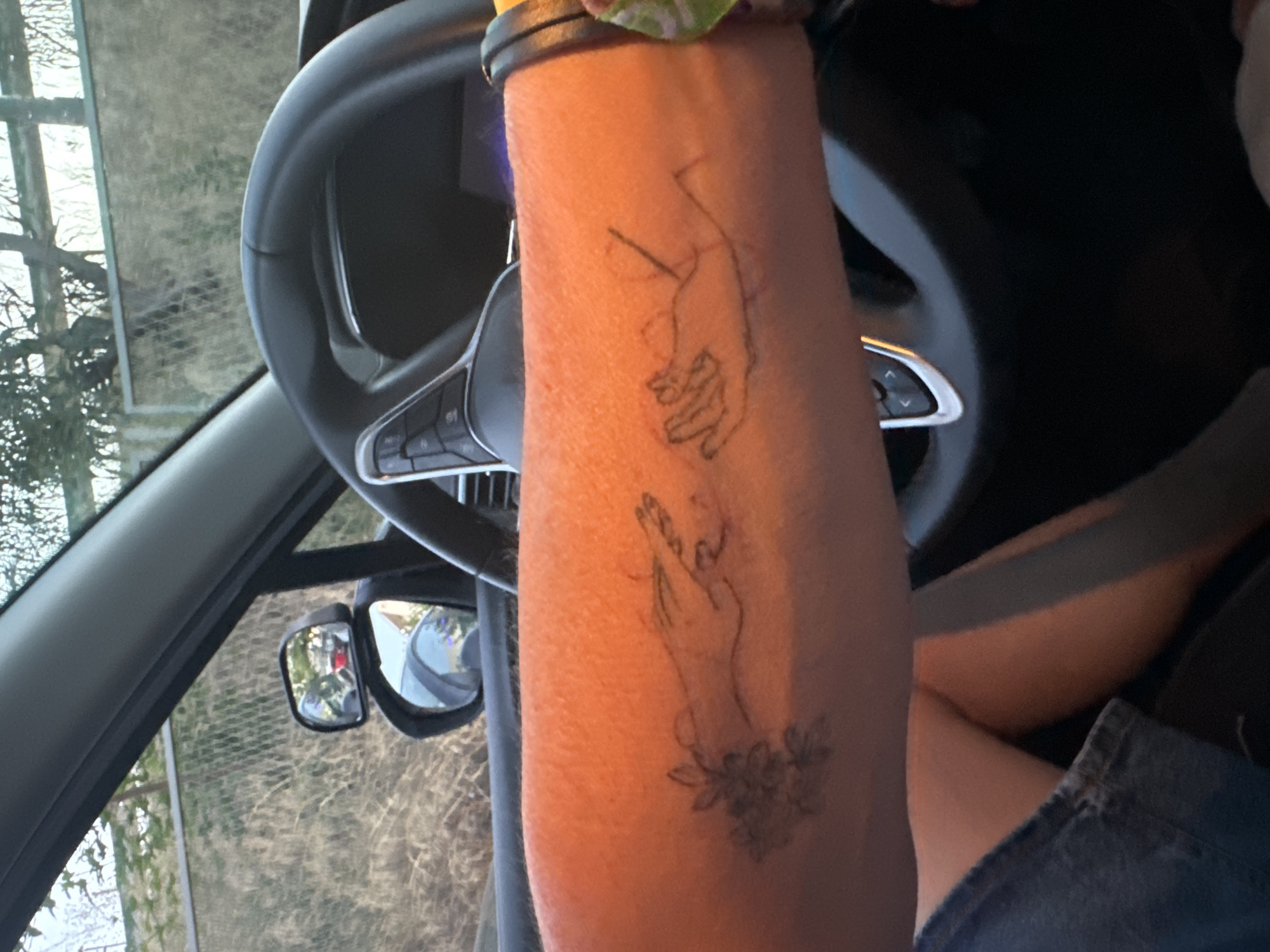 An outdoor daytime medium shot shows a person's tattooed forearm and hand resting on the steering wheel of a car, with the steering wheel and interior of the car visible in the foreground and midground. The person's arm, tattooed with line drawings of two hands reaching for each other and a cluster of flowers, is illuminated by the sun. The steering wheel is black and features several control buttons. A car's side mirror is visible in the background, reflecting the surrounding environment. The scene appears to be set in a rural or suburban area, with dry vegetation and a fence visible through the car's windows. The image does not provide any information about the city of Murcia, Spain.