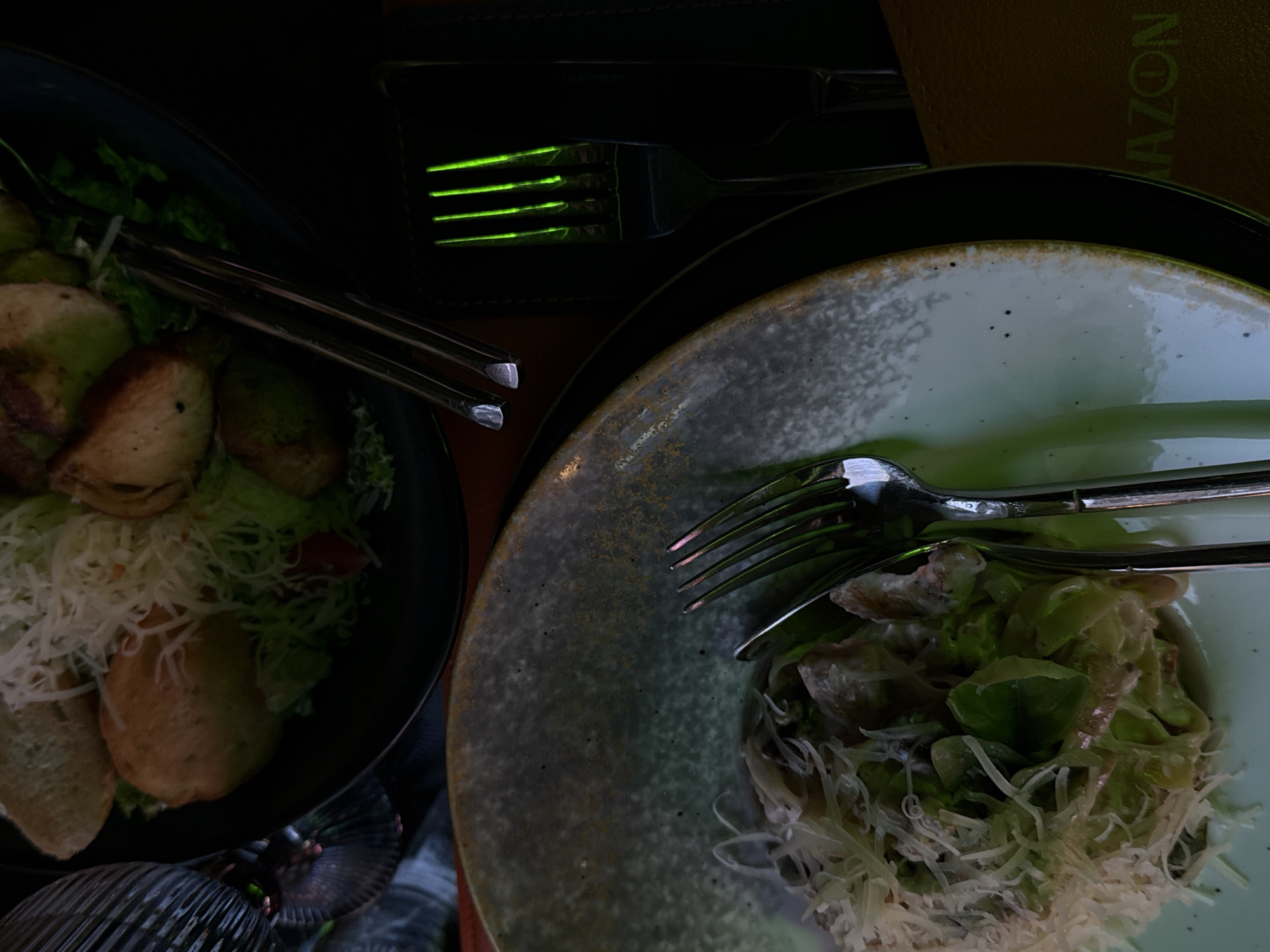 The image shows two plates of salad. One plate contains a Caesar salad with croutons, lettuce, shredded cheese, and dressing. The other plate contains a salad with lettuce, shredded cheese, and dressing. Silverware is placed on both plates. There is a green glow reflected in the forks in the background. The image was likely taken in Yerevan, Armenia.