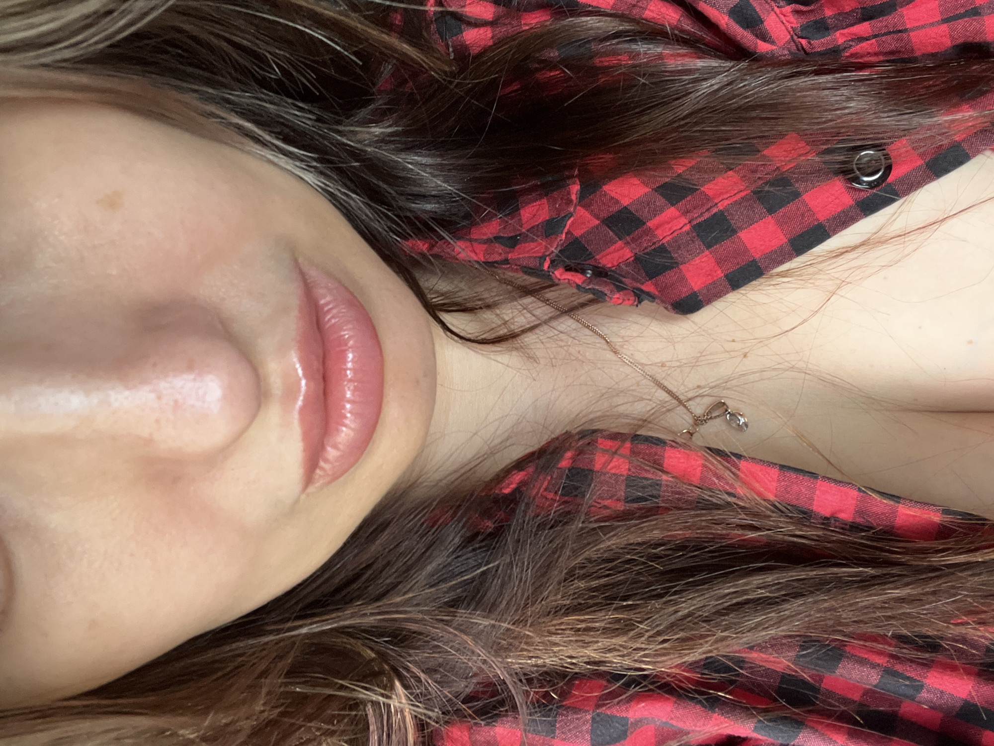 A close-up, chest-level portrait of a person features the lower portion of the face, neck, and upper chest. The individual has long, dark brown hair with lighter strands, which frames the face and falls over the shoulders and a red and black plaid shirt. The visible facial features include a portion of the nose, exhibiting some natural texture and slight redness on the tip, and full, glossy, light pink lips that are slightly parted. The skin is light-toned, with visible pores and minor marks, including a small brown nevus on the upper right cheek area. A red and black buffalo check button-up shirt is worn, unbuttoned to reveal the upper sternum and décolletage. A thin, metallic chain necklace with a small, reflective charm is visible, resting on the chest. The charm appears to be a stylized "C" or crescent shape with a small, clear embellishment. A single black button is discernible on the right side of the open shirt. The environment is entirely obscured by the close-up perspective of the subject. The location in the city of Unknown, Unknown, is not discernible from this image.