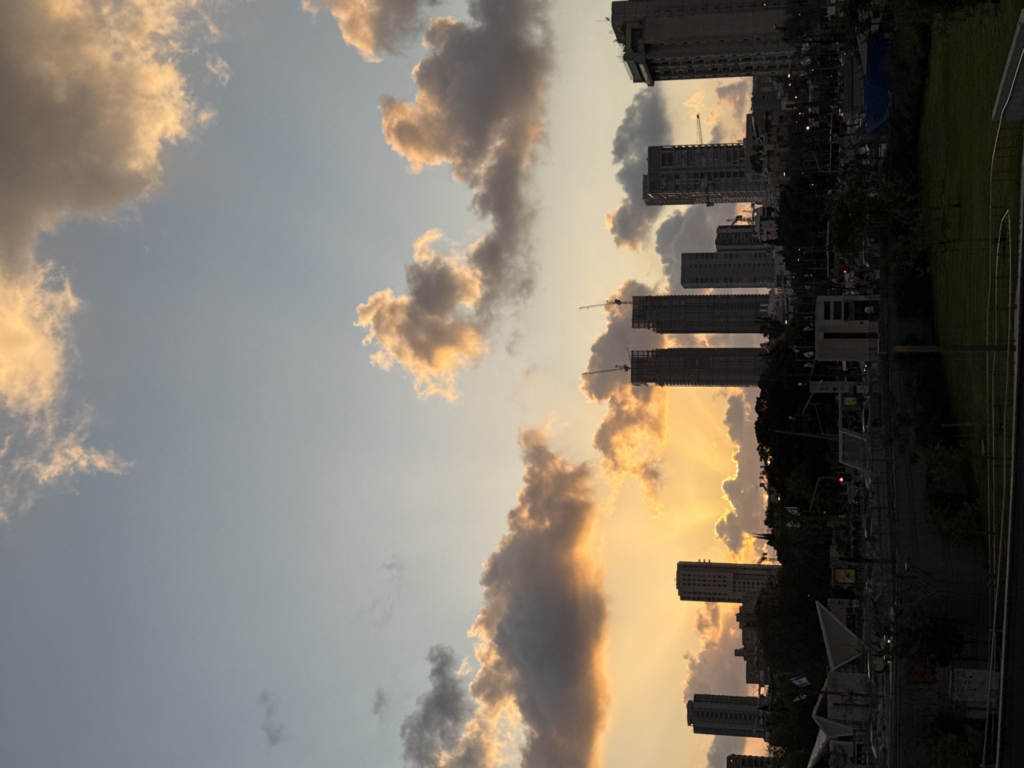 The image displays a panoramic view of a city skyline at either sunrise or sunset, set against a dramatically lit sky. The scene is likely Tel-Aviv, Israel, as per the provided context. The sky is a prominent feature, transitioning from a soft blue at the top to a brilliant orange and golden yellow near the horizon, indicating the low angle of the sun. Scattered across the sky are numerous clouds, some fluffy and brightly illuminated by the golden light, while others appear darker and more shadowed, adding depth and contrast.
Beneath the radiant sky, the cityscape is largely silhouetted. A series of tall, modern buildings define the urban horizon, some appearing complete while others feature prominent construction cranes, suggesting ongoing development. The buildings are dark against the bright sky, with minimal detail visible in their shadowy facades. In the foreground, below the buildings, hints of urban infrastructure are present, including what appears to be a street or pathway, and some dark trees and grassy areas, all cast in deep shadow. The overall atmosphere is serene and striking, characterized by the warm, dramatic lighting of dusk or dawn. No discernible text is visible in the image.