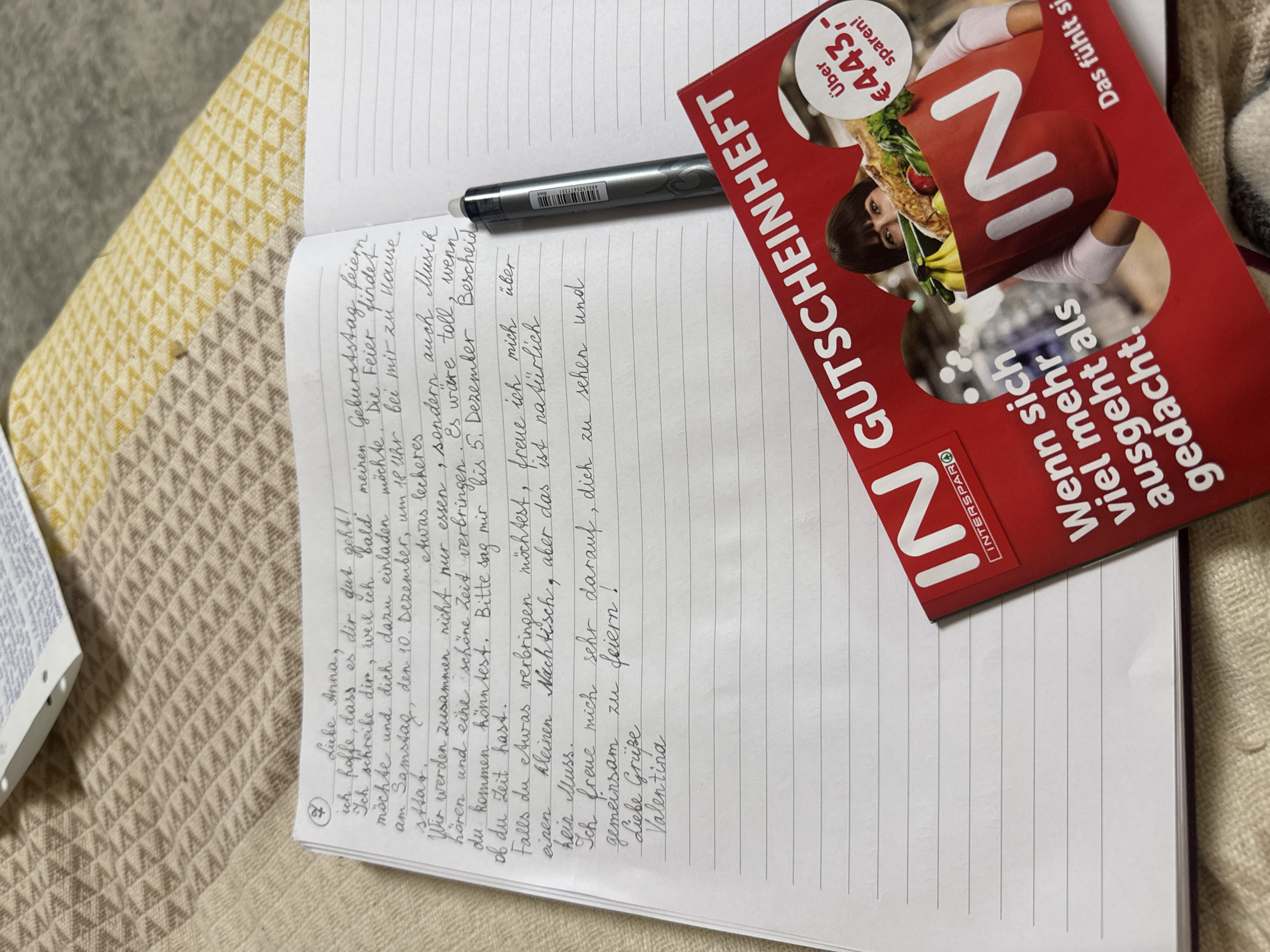 The image shows a notebook with handwritten German text, a pen, and a red coupon booklet. The notebook is open to a page with a letter addressed to "Liebe Anna". The letter discusses plans for a birthday celebration on Saturday, December 10th, at 6 PM, at the sender's home, including food and music. The sender asks Anna to confirm her attendance by December 5th. The letter is signed "Valentina". The coupon booklet is red and features the word "GUTSCHEINHEFT" and an image of a woman holding a shopping bag. The text on the booklet includes "IN" and "INTERSPAR", and a mention of saving "€443,-". The notebook and coupon booklet are placed on a textured surface, likely a blanket or fabric. There is no visible information indicating the specific location within Sankt Andrä-Wördern, Austria.