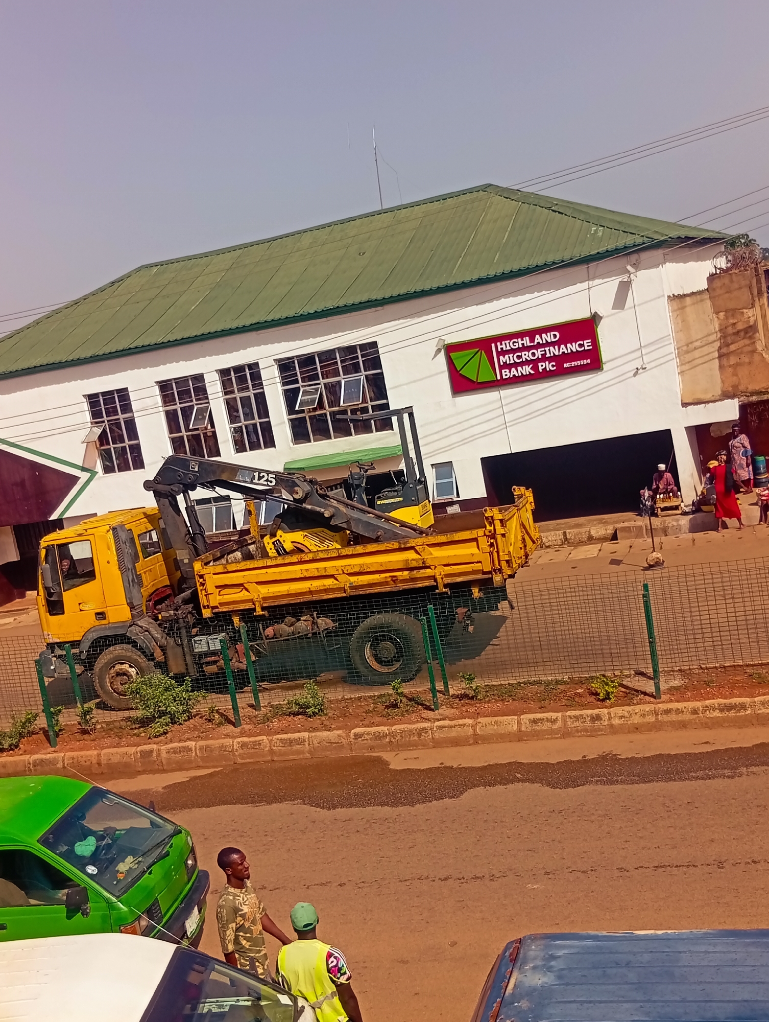 The image depicts a street scene in Bukuru, Nigeria, with a building labeled "HIGHLAND MICROFINANCE BANK PLC" in the background. In front of the bank, a yellow flatbed truck with a crane is parked on the street. The truck is a commercial vehicle, likely used for construction or transport. The building features a green roof and multiple windows. Several people are visible near the entrance of the bank, some appearing to be customers or vendors. In the foreground, two men are standing near a green van, suggesting an interaction or waiting. Another vehicle, a portion of a white vehicle, is visible to the left. The street is paved, and a low metal fence separates the sidewalk from the road. The environment appears to be a commercial or semi-urban area within Bukuru. The overall lighting suggests it is daytime with clear weather.