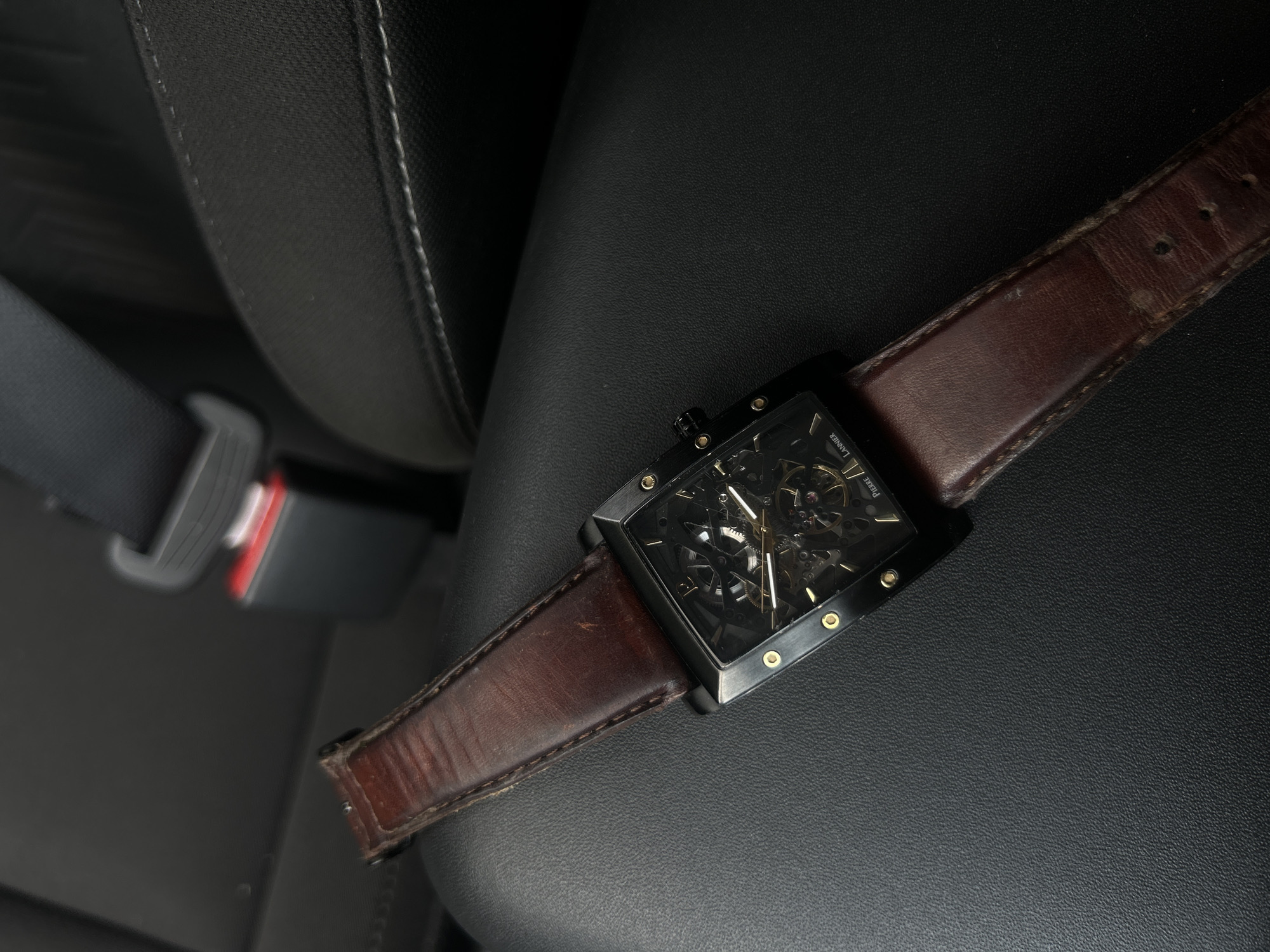 A close-up shot captures a stylish watch resting on a black, textured surface, likely a car seat. The watch features a rectangular black case with prominent gold-colored screws or markers on its edges. Its most striking detail is the skeletal dial, which reveals the intricate gears and mechanisms of the watch movement beneath the hands. The watch is fitted with a dark brown leather strap, showing visible signs of wear, creases, and minor scuffs, indicating its age and use.
In the blurred background, to the upper left, a car seatbelt buckle with red and silver accents is visible, confirming the setting as a vehicle's interior. The black surface beneath the watch has a subtle, pebbled texture. The lighting appears to be soft, creating gentle reflections on the watch case and highlighting the texture of the seat.
Visible text on the watch dial, near the right edge, reads "PIERRE" stacked above "LANNIER", identifying the brand as Pierre Lannier. There are no people visible, and no specific activity is taking place other than the watch resting in the car. The image is a static portrayal of an accessory in a common everyday setting.